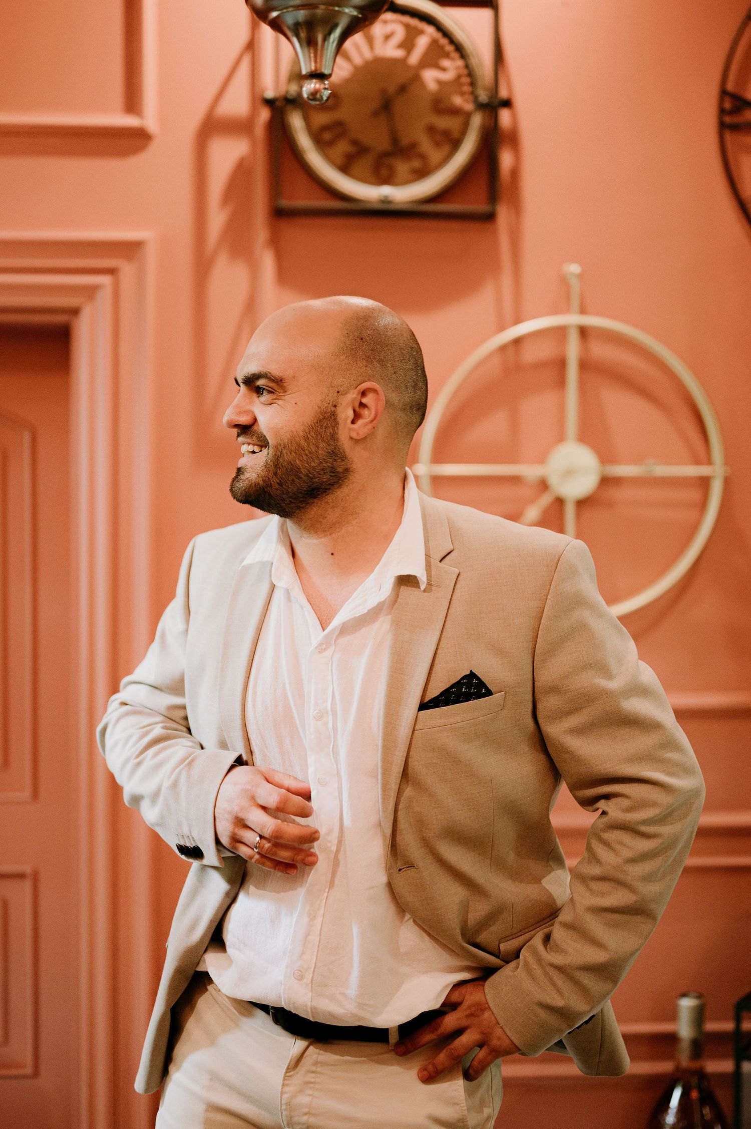 A person in a beige suit poses against a coral-colored wall with decorative moldings.