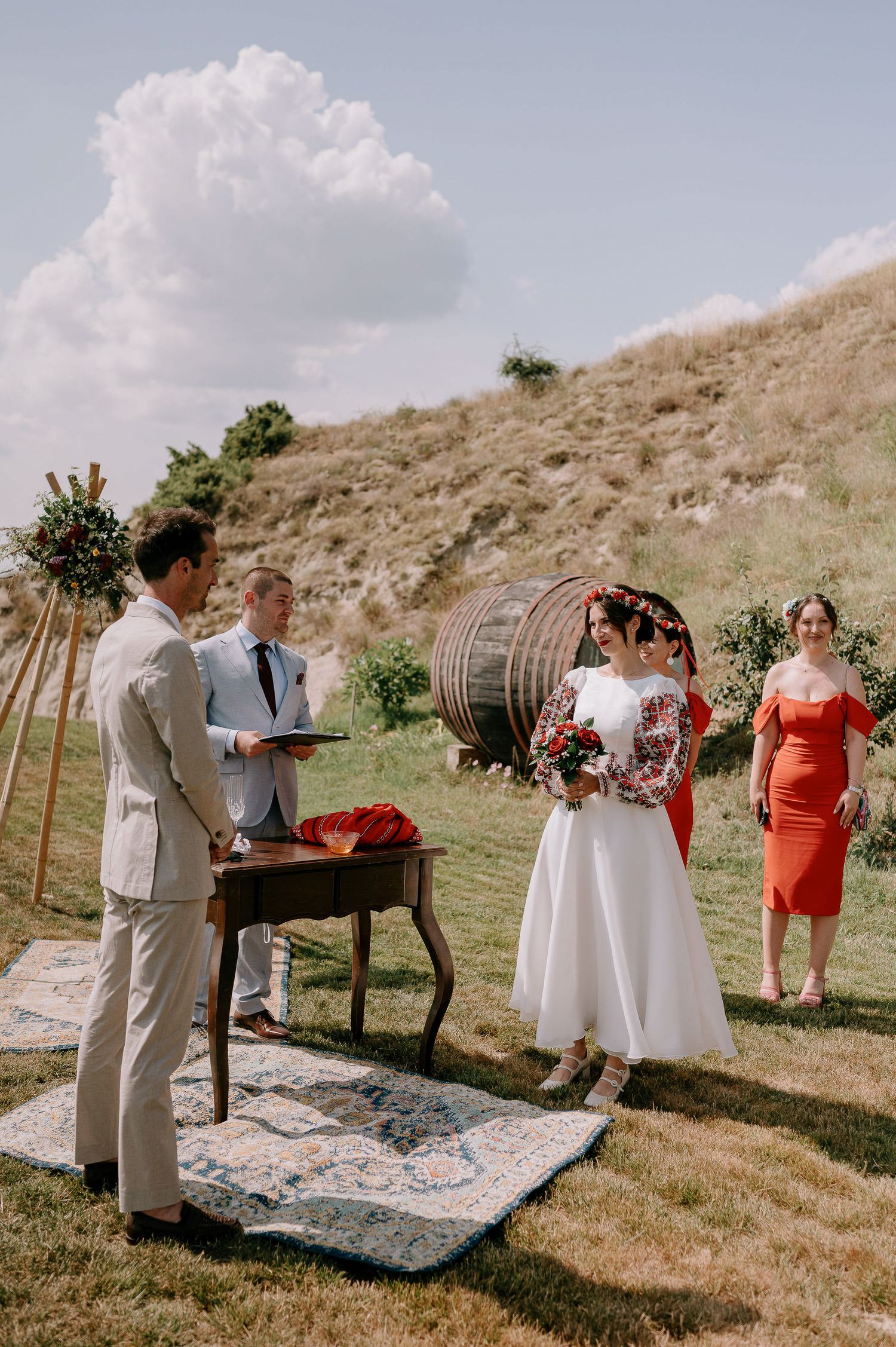 Wedding ceremony taking place on a hillside with wine barrels and decorative rugs.