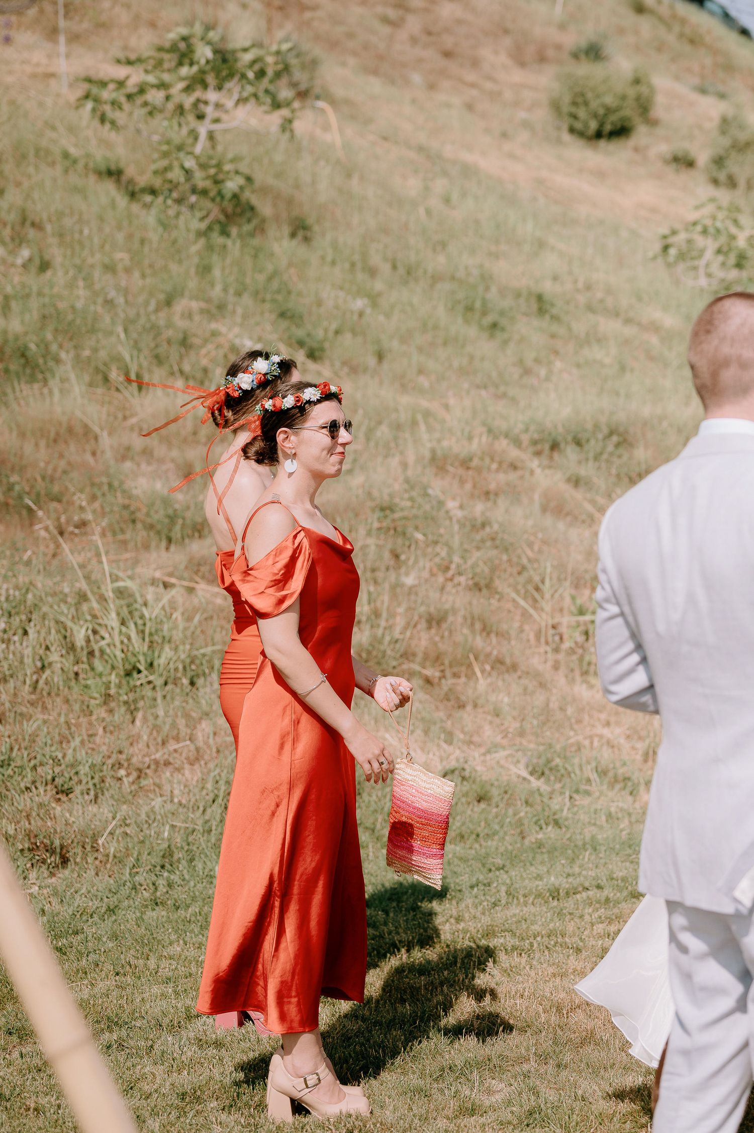 Wedding guests in coral dresses and light suits mingle on sunny hillside.