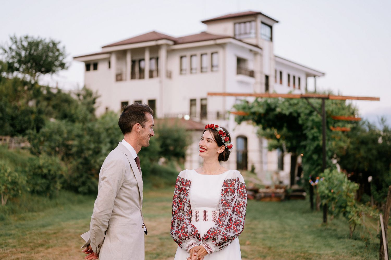 A wedding couple poses outdoors near a Mediterranean-style building in vintage-inspired attire.