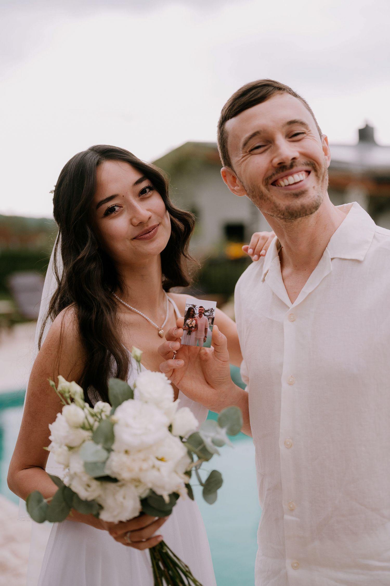 A couple in white outfits posing together by a pool with flowers.