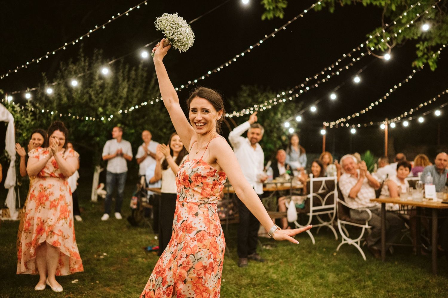 Guest in coral dress celebrates joyfully at outdoor wedding reception under string lights.