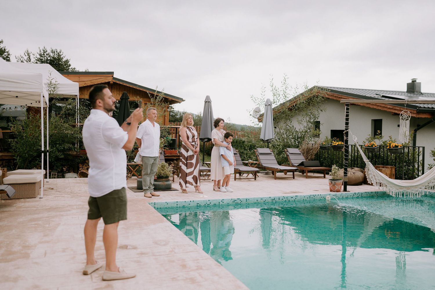 Casual outdoor celebration by a swimming pool with wooden deck and pergola.
