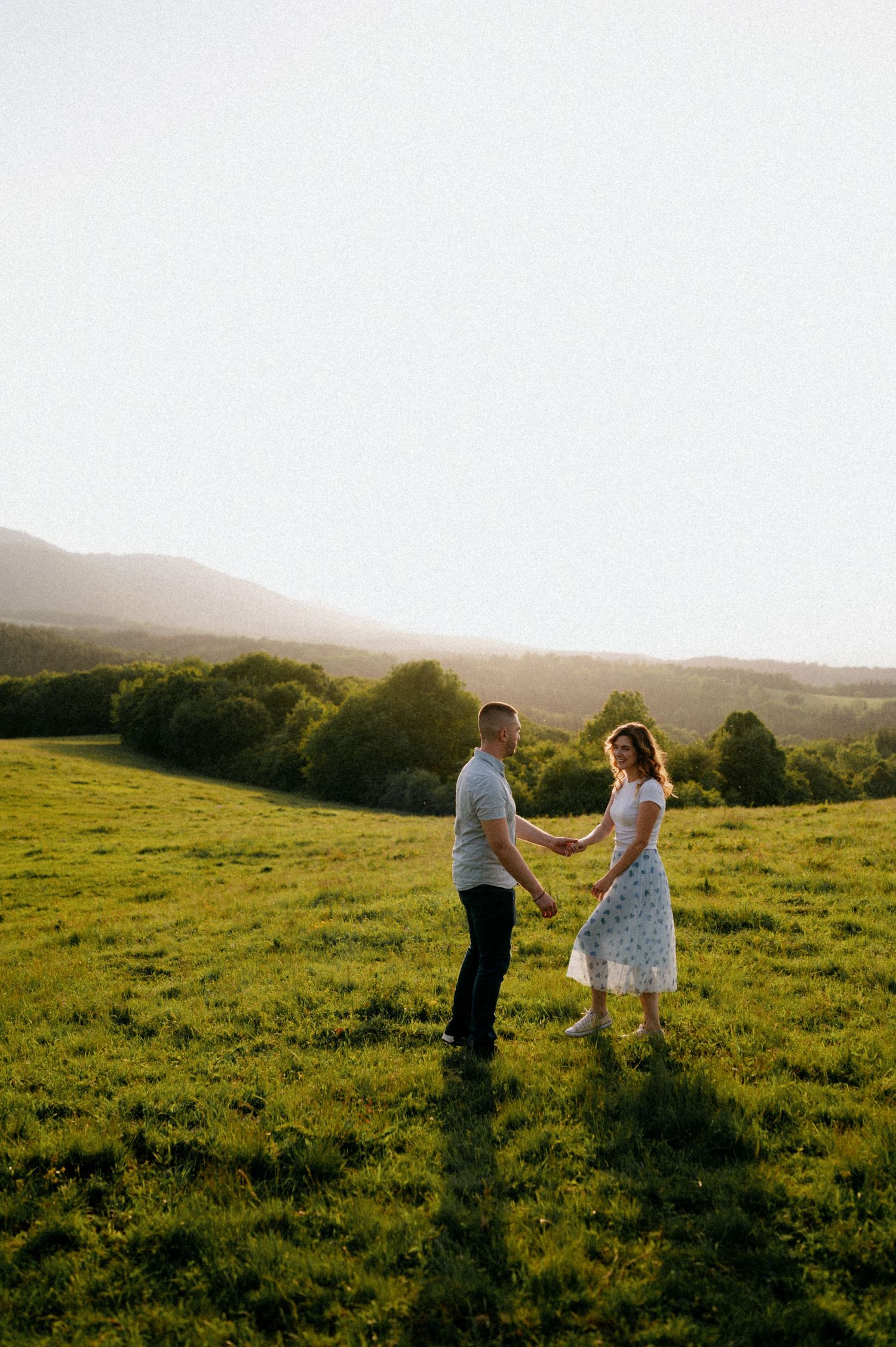 A couple dances together in a scenic green meadow at sunset with mountains in the background.
