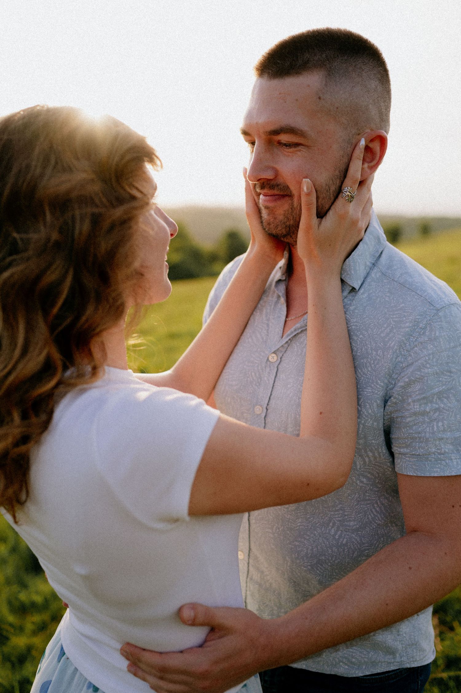 A couple laughs together while holding each other's faces in a green meadow at sunset.