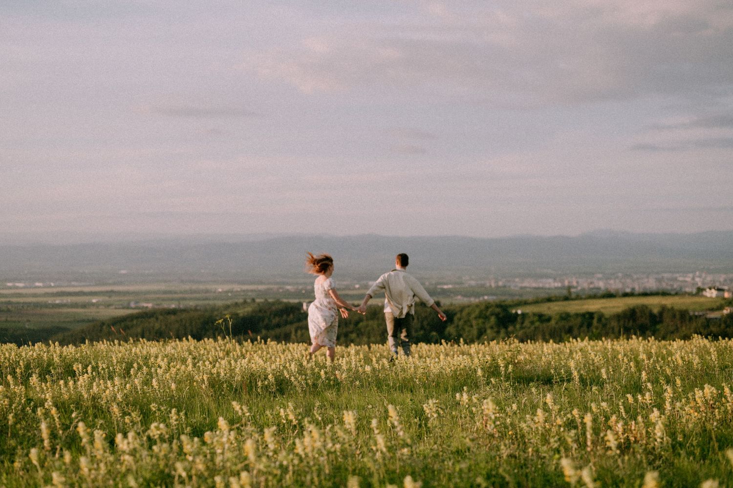 Two figures hold hands while walking through a golden meadow with mountain views.