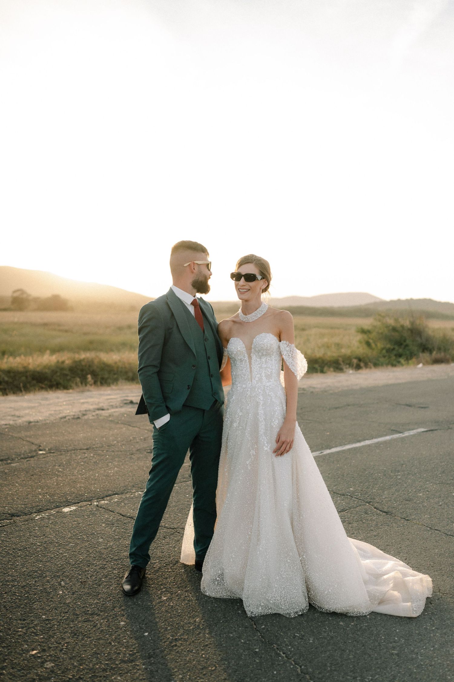 Bride in lace gown and groom in teal suit pose on road at golden hour with rolling hills backdrop.