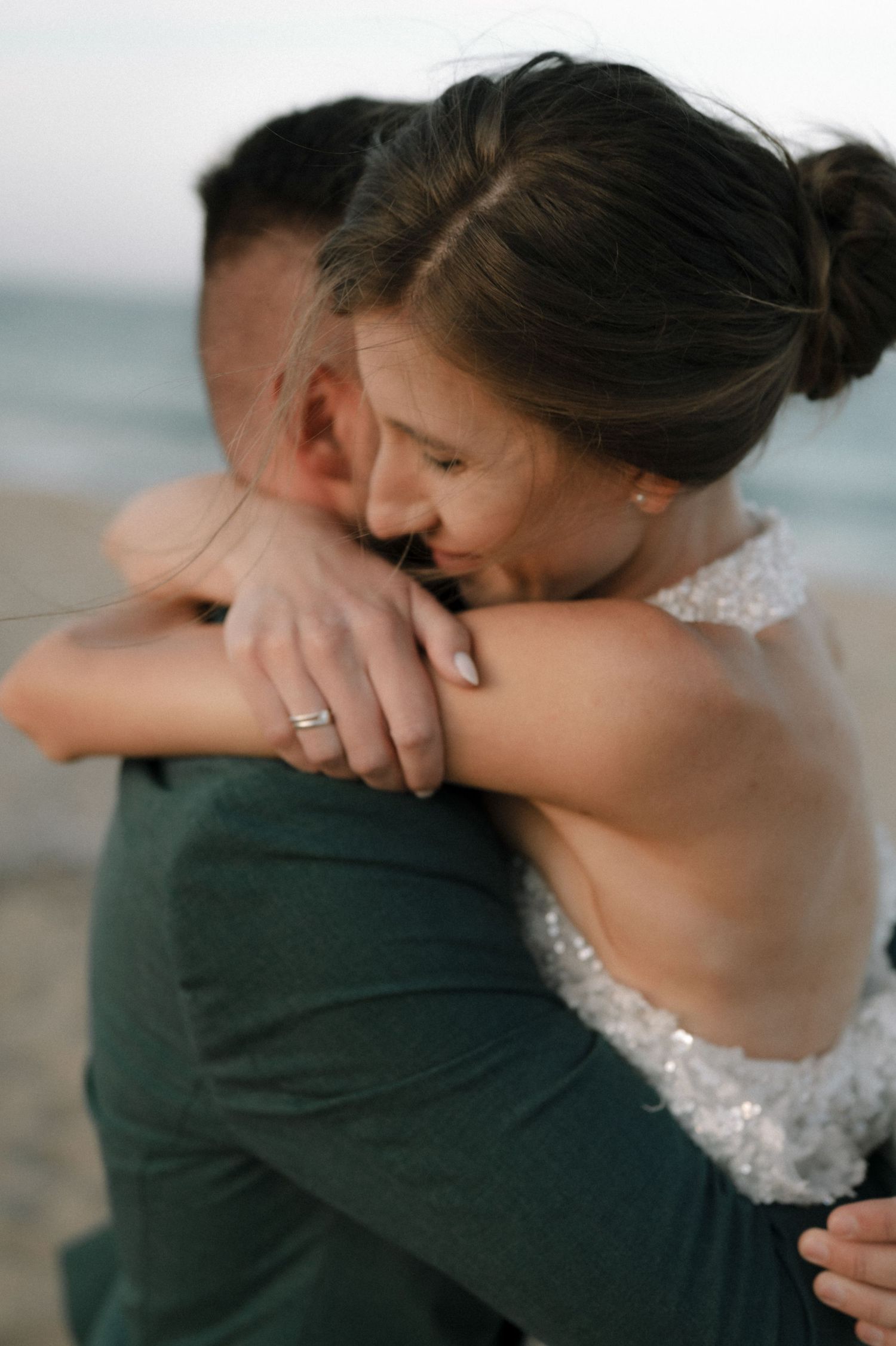 A bride in a lace gown embraces her groom on a beach, smiling with joy, wearing a diamond ring.