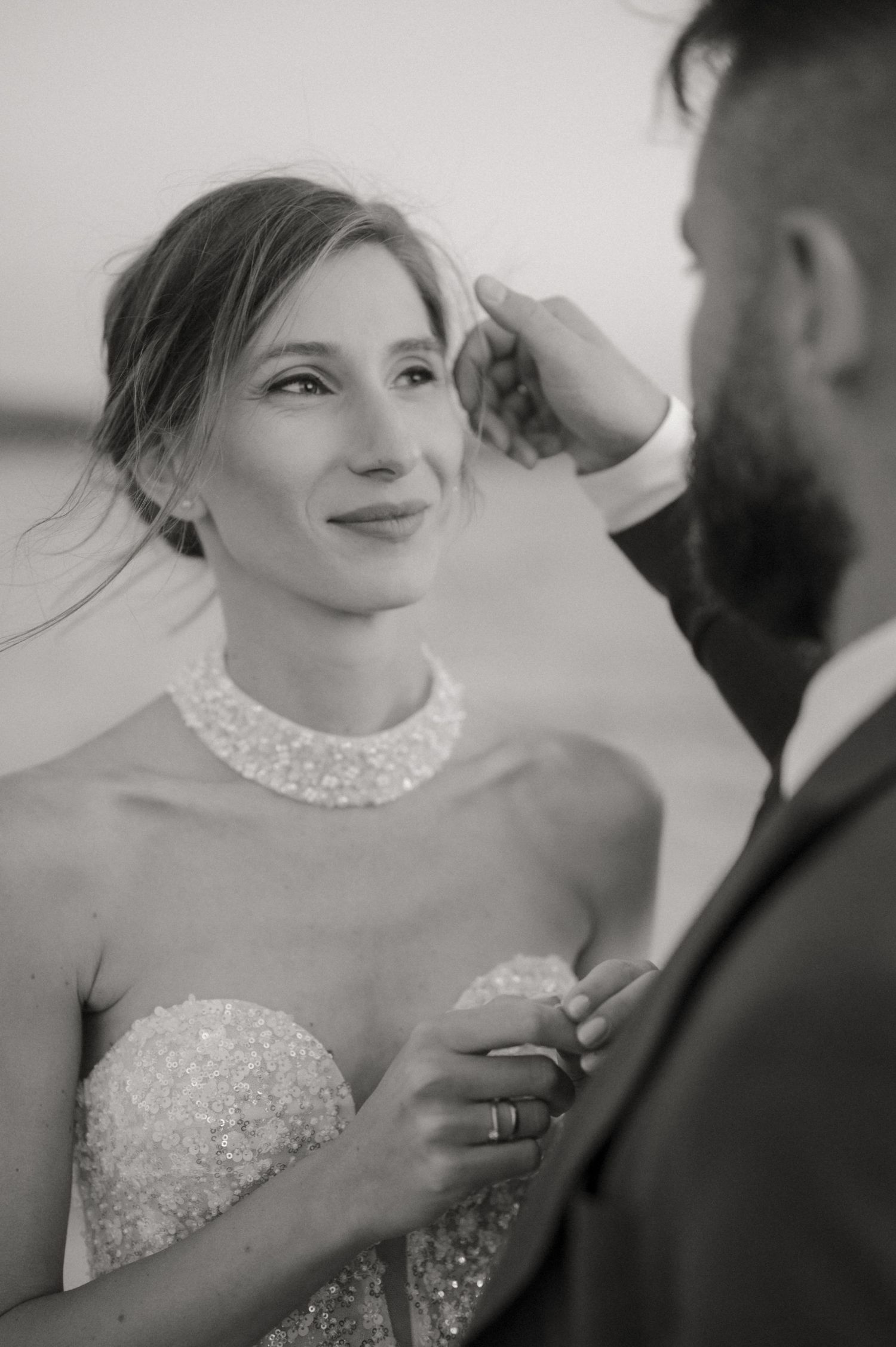 Black and white wedding photo of bride in beaded gown with pearl necklace, smiling as groom gently touches her face.