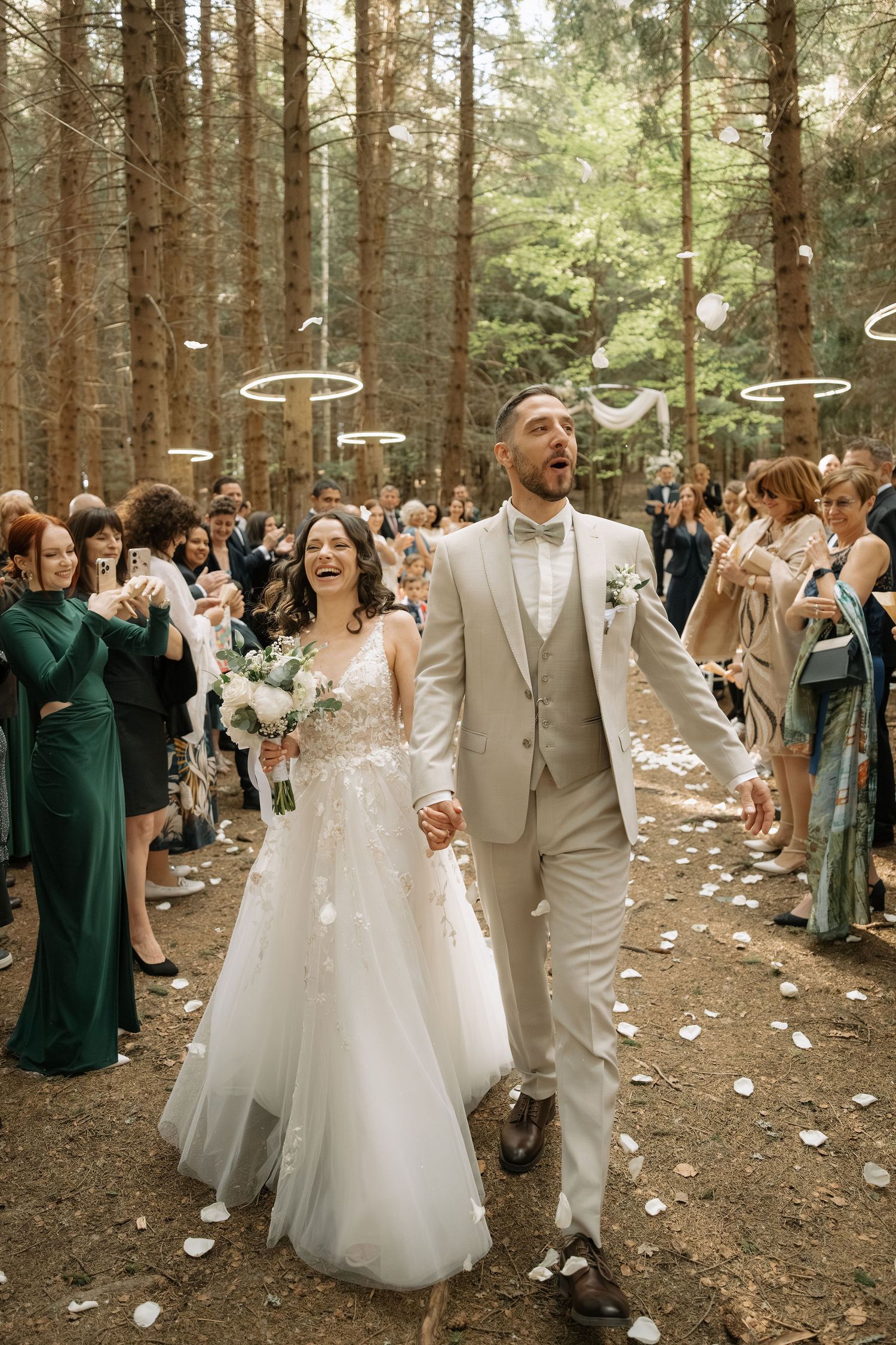 Joyful bride and groom walk through forest aisle as guests toss white petals during outdoor wedding ceremony.