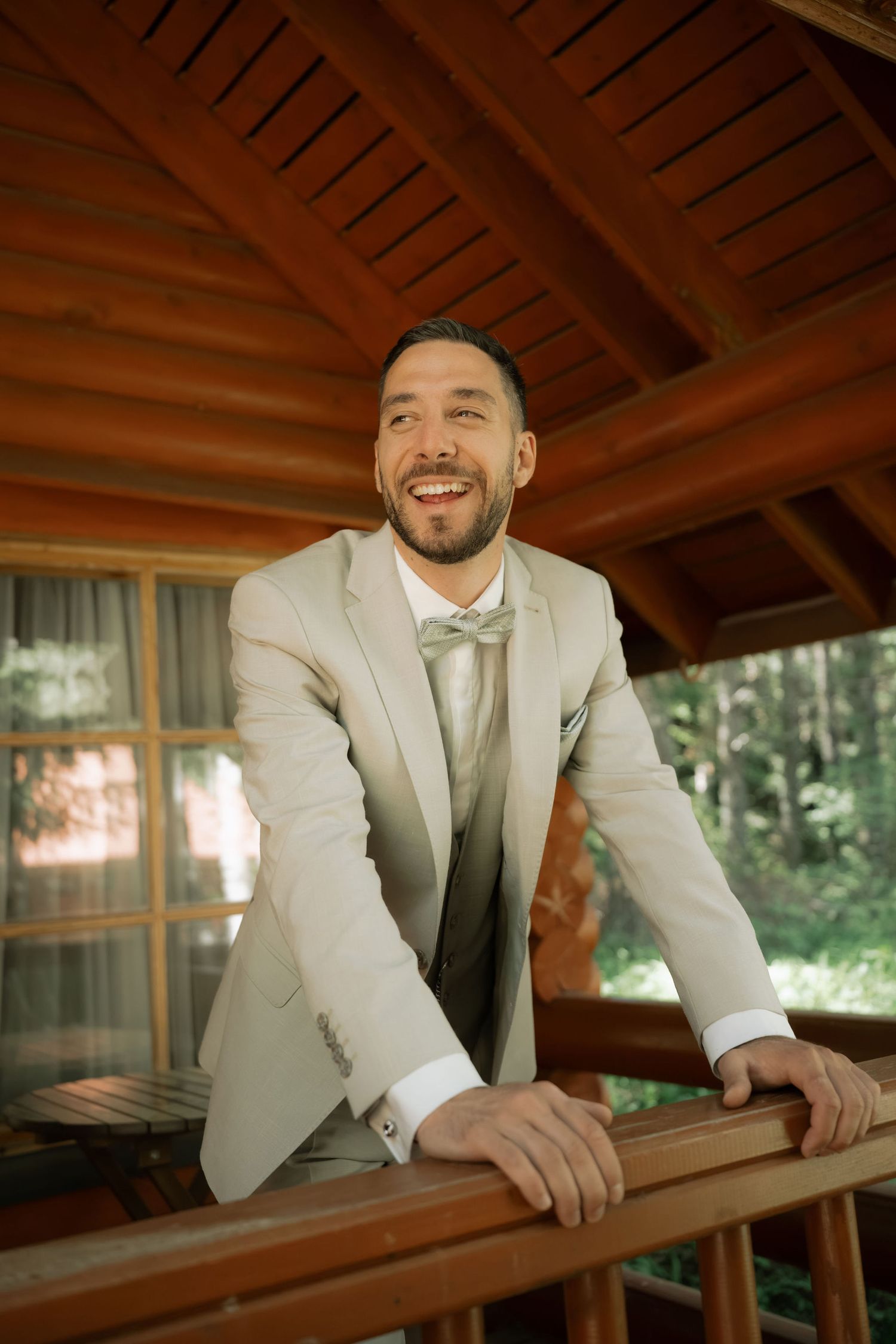 Smiling groom in beige suit and bow tie leaning on wooden railing of rustic cabin balcony surrounded by forest.