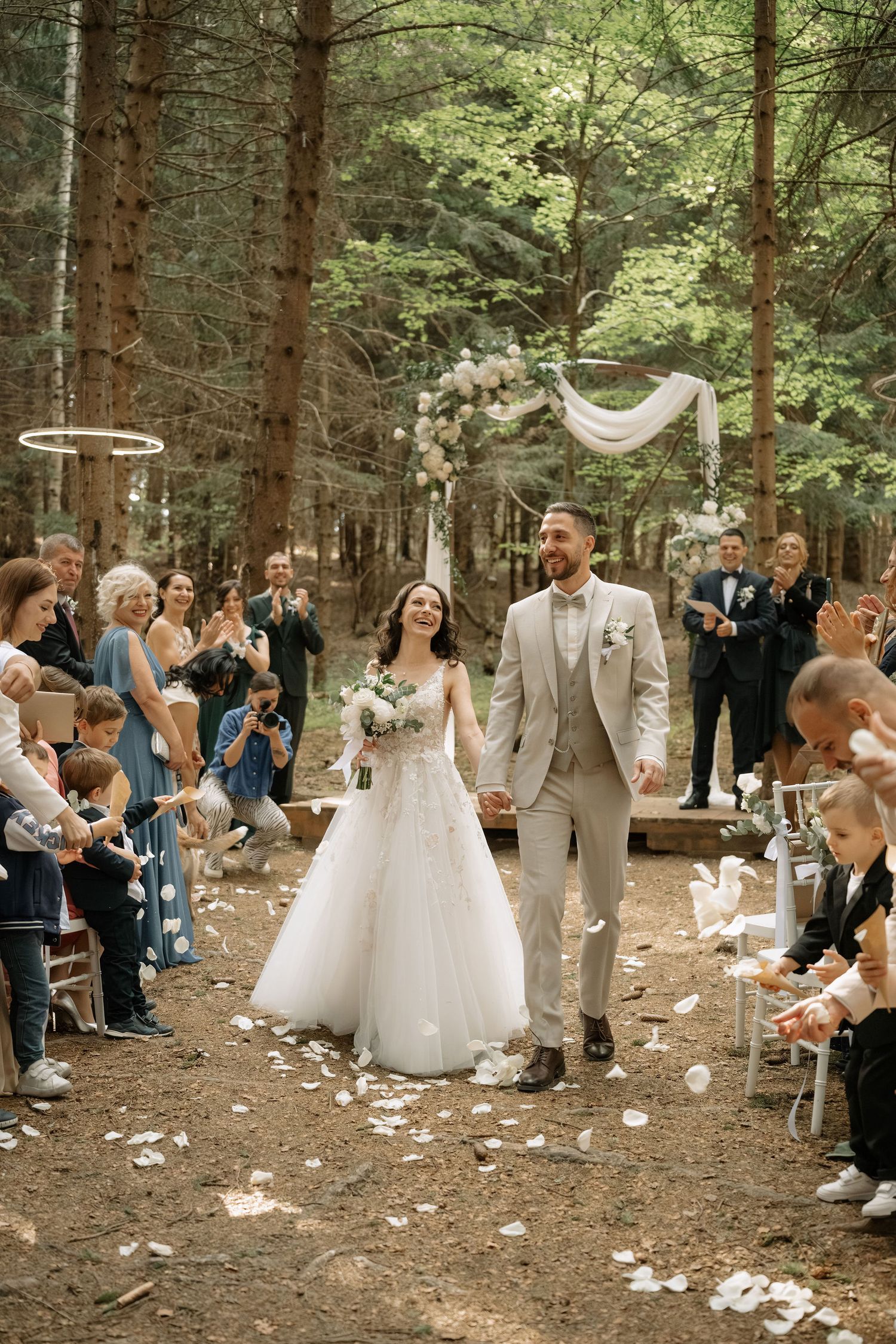 Bride and groom walk down forest aisle as guests toss flower petals at outdoor woodland wedding ceremony.