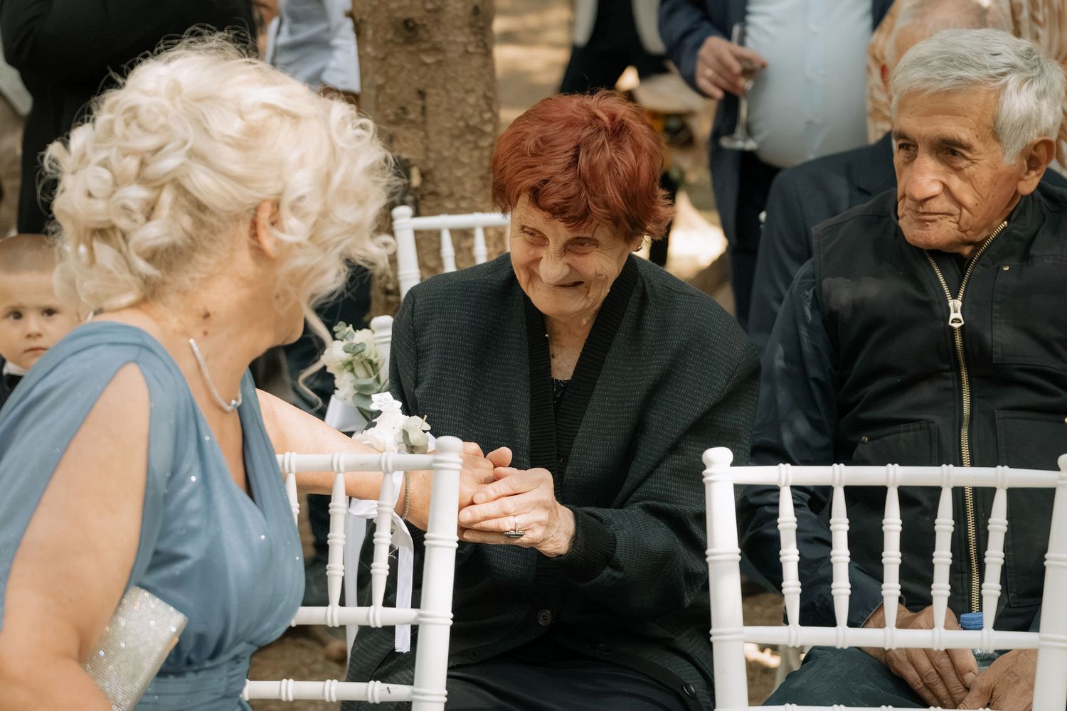 Elderly guests chat and hold hands at an outdoor wedding ceremony seated on white chairs.