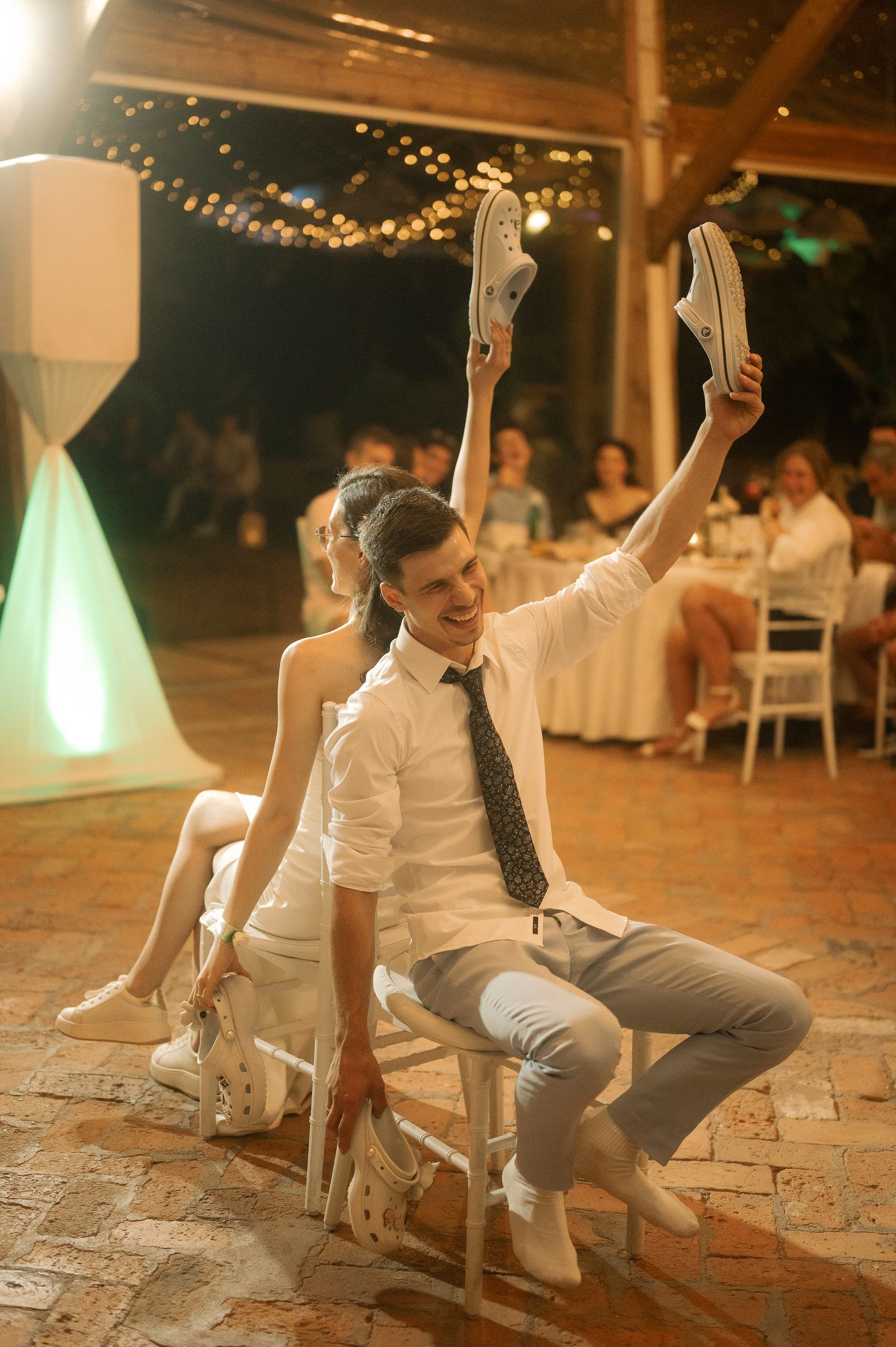 Two wedding guests sit in chairs back-to-back during a fun reception game on a dance floor with string lights overhead.