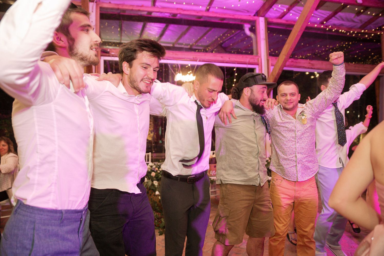 Energetic group dancing in formation during a wedding reception with pink and purple ambient lighting.