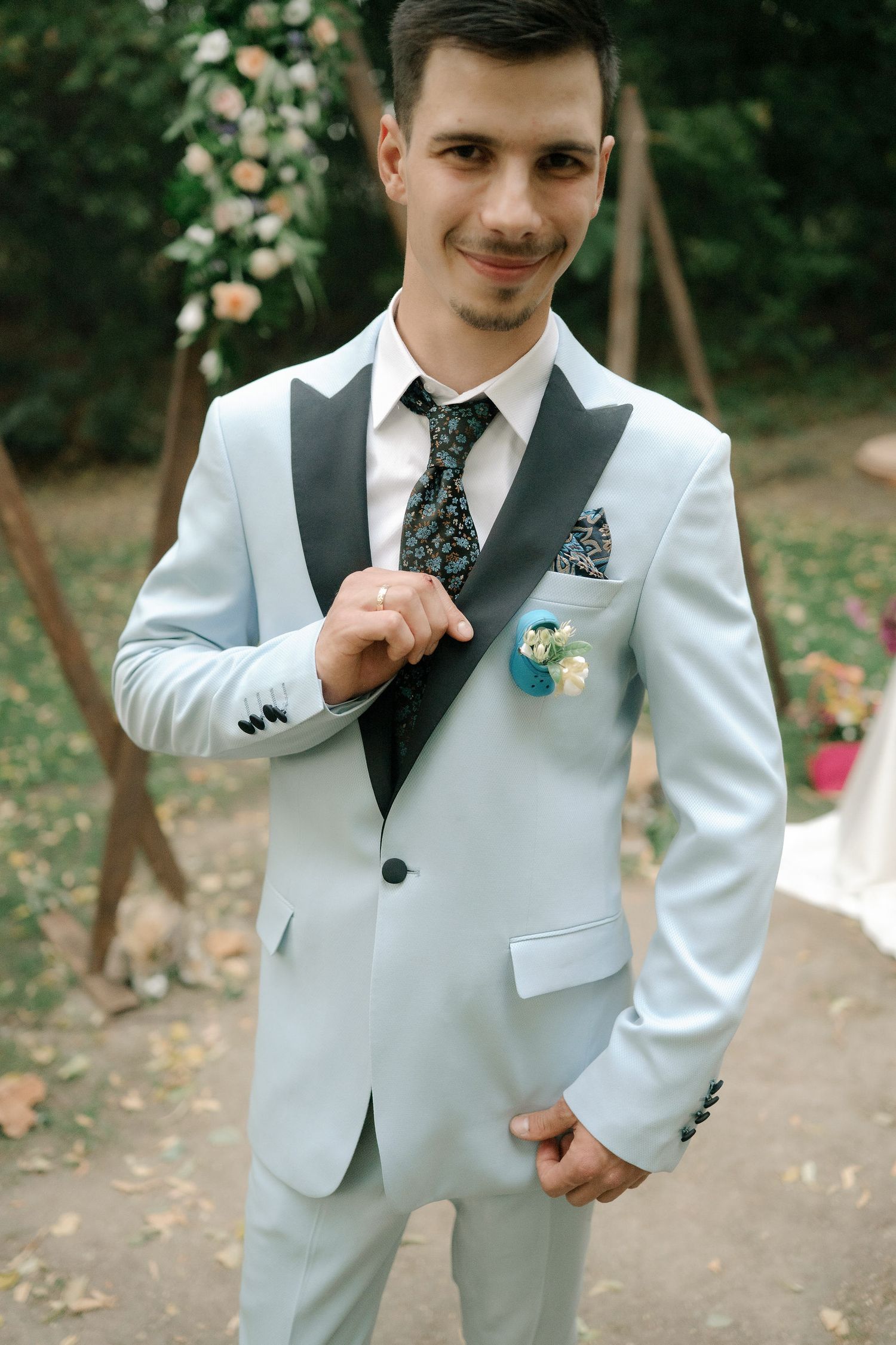 A formal white suit with a floral tie and boutonniere is worn at an outdoor garden event.