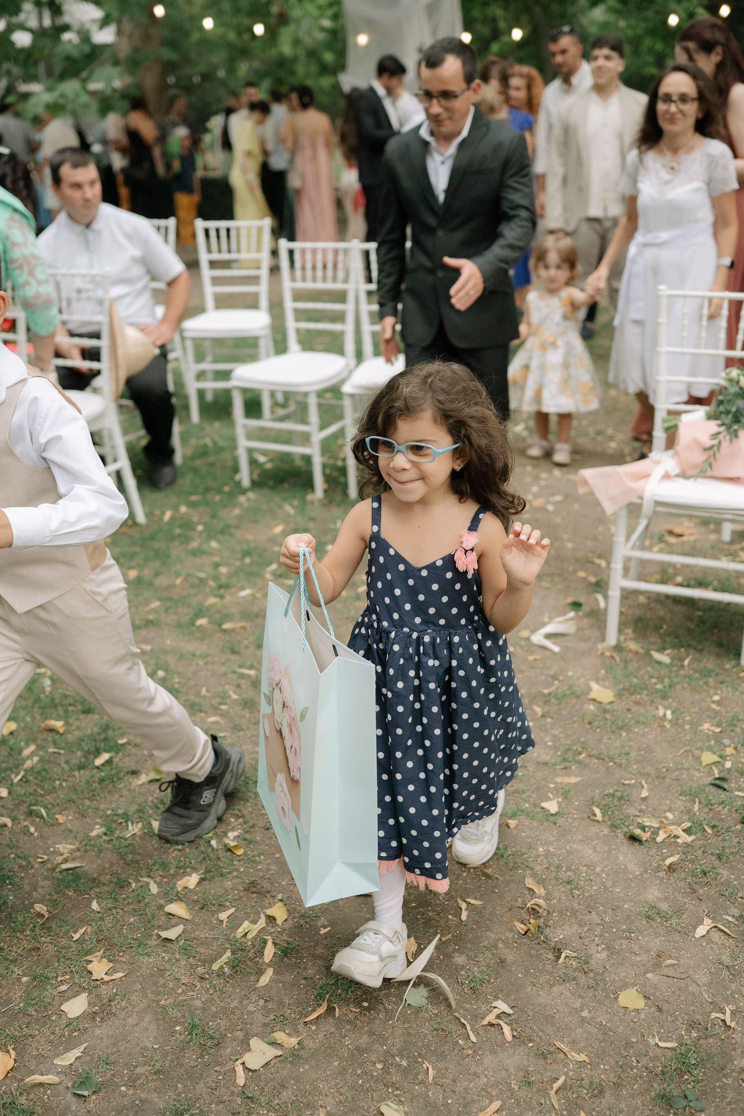 A young child in a polka dot dress dances and twirls joyfully at an outdoor garden party with white chairs.