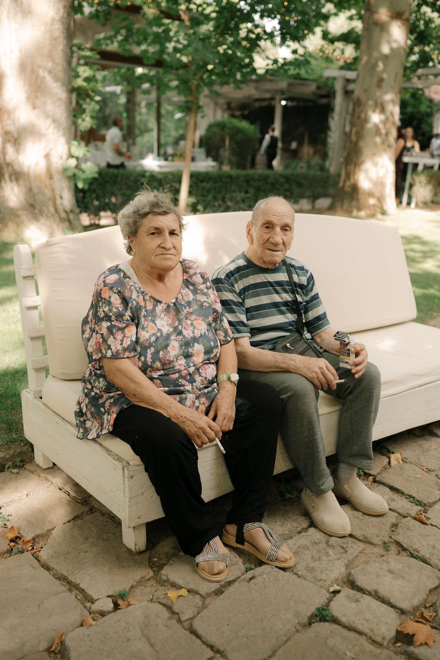 An elderly couple sits together on a light-colored outdoor bench surrounded by fallen autumn leaves.