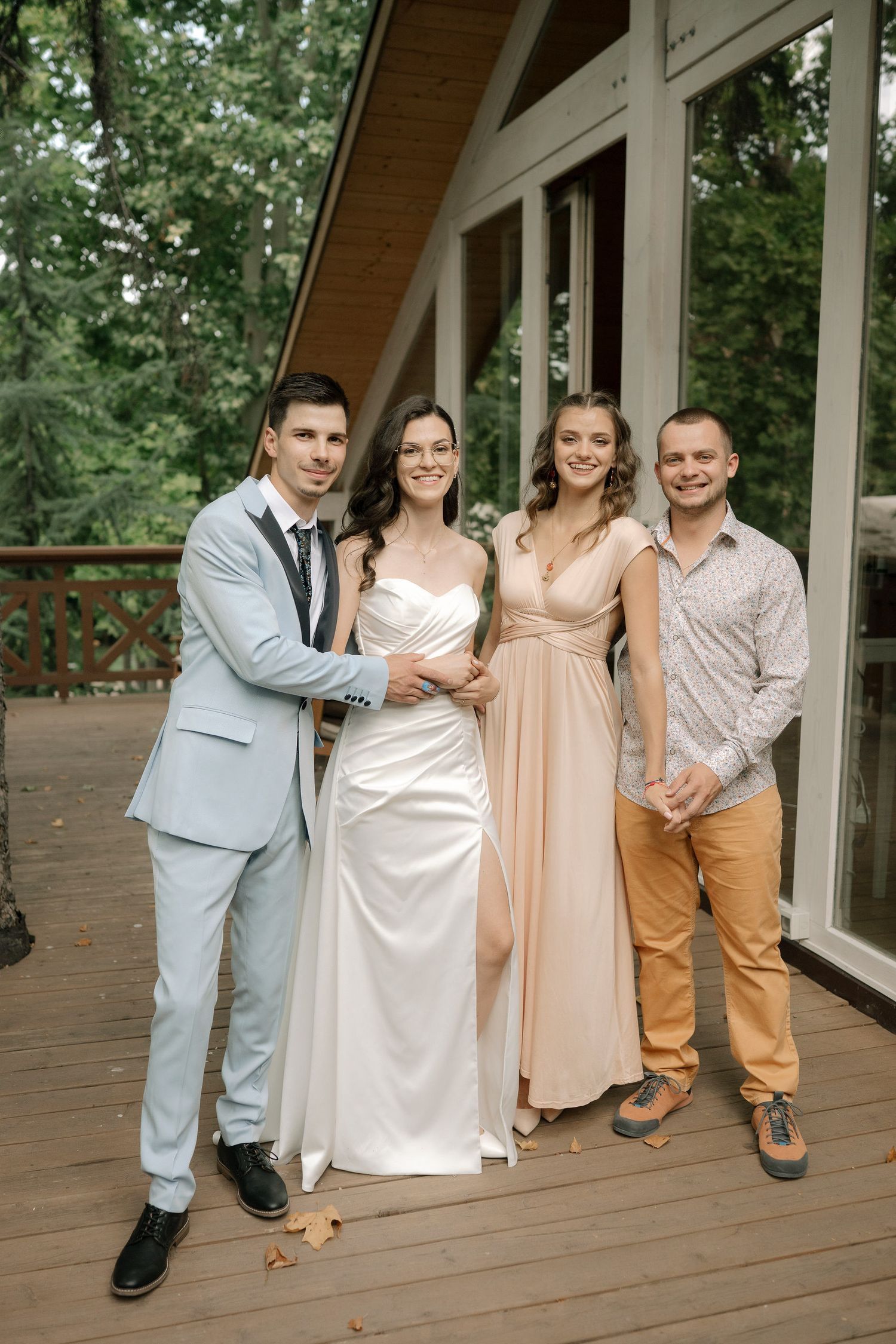 A wedding party poses on a wooden deck in front of a modern A-frame building surrounded by trees.