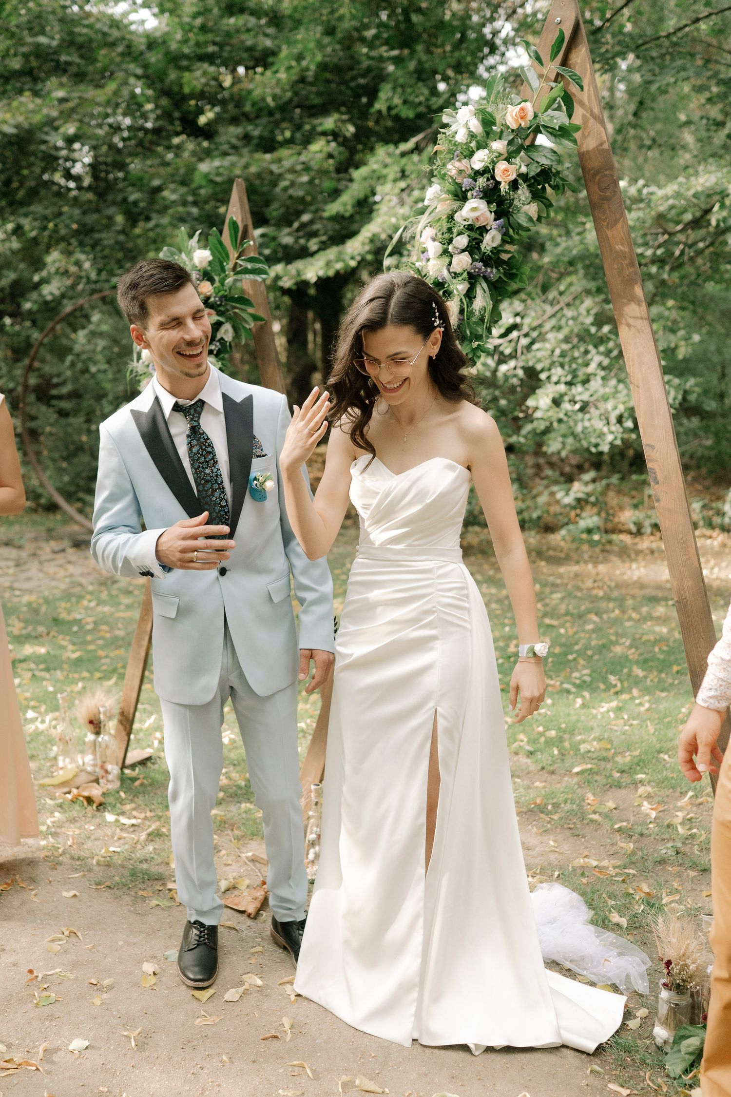 A couple in wedding attire laughs together while walking through a wooded outdoor setting.