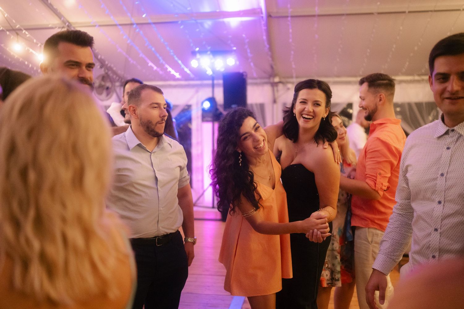 Wedding guests dancing and celebrating on a dance floor illuminated with purple lighting at an indoor reception.
