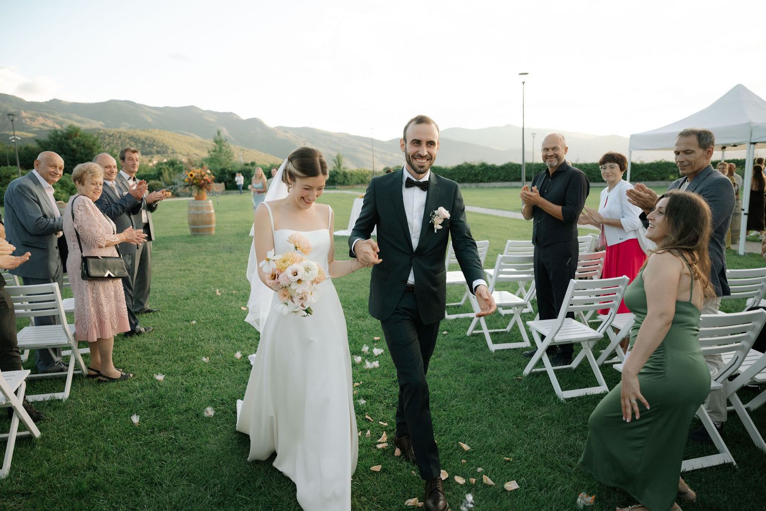 Outdoor wedding ceremony guests mingle on a grassy lawn with mountain views during golden hour.