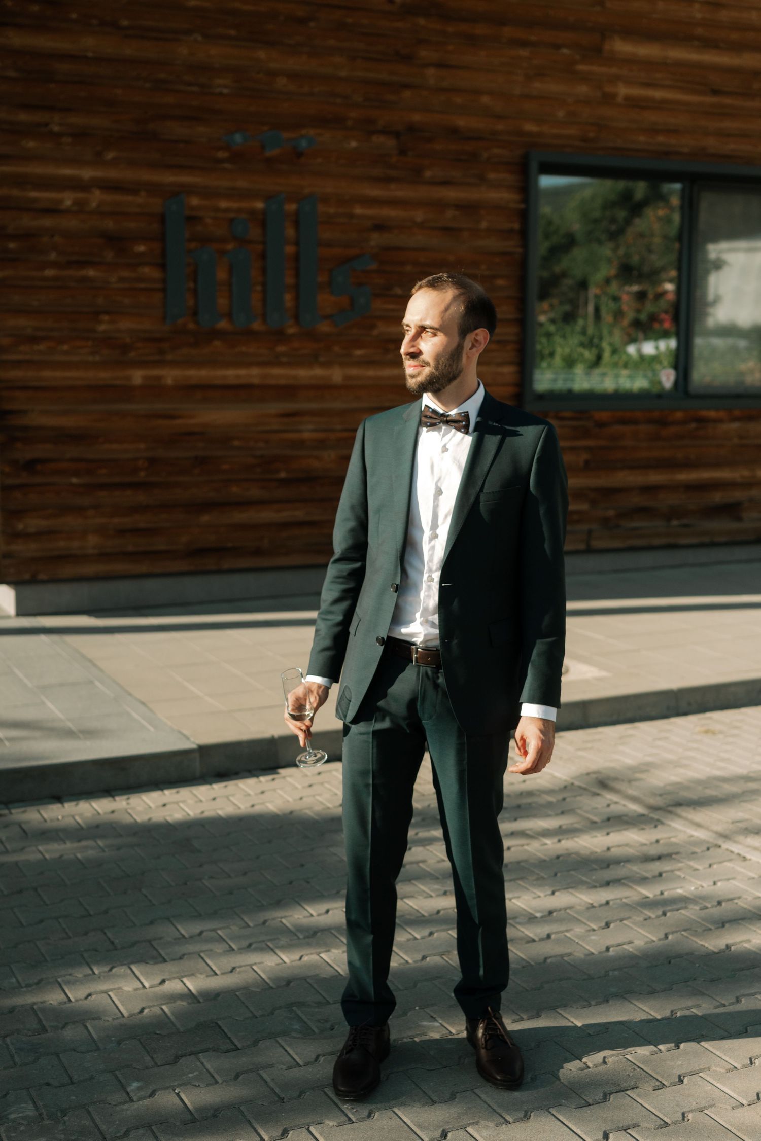 A person in a formal black tuxedo and bowtie stands on a paved walkway outside a wooden-paneled building.