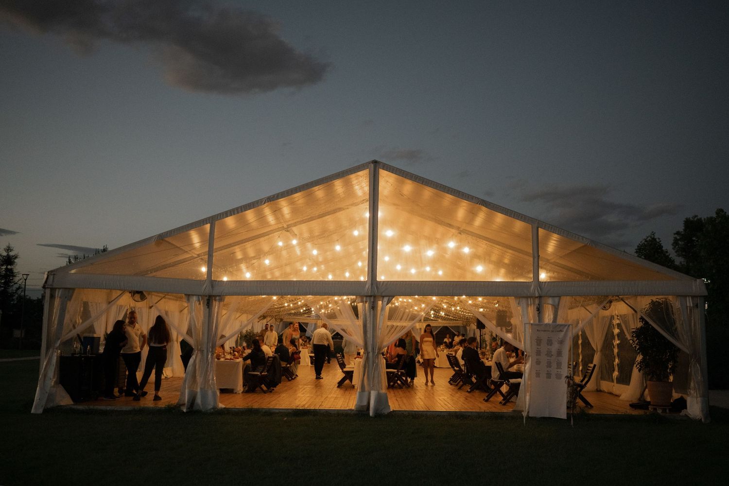 A row of illuminated white wedding tents glowing at dusk with string lights creating a warm, romantic atmosphere.