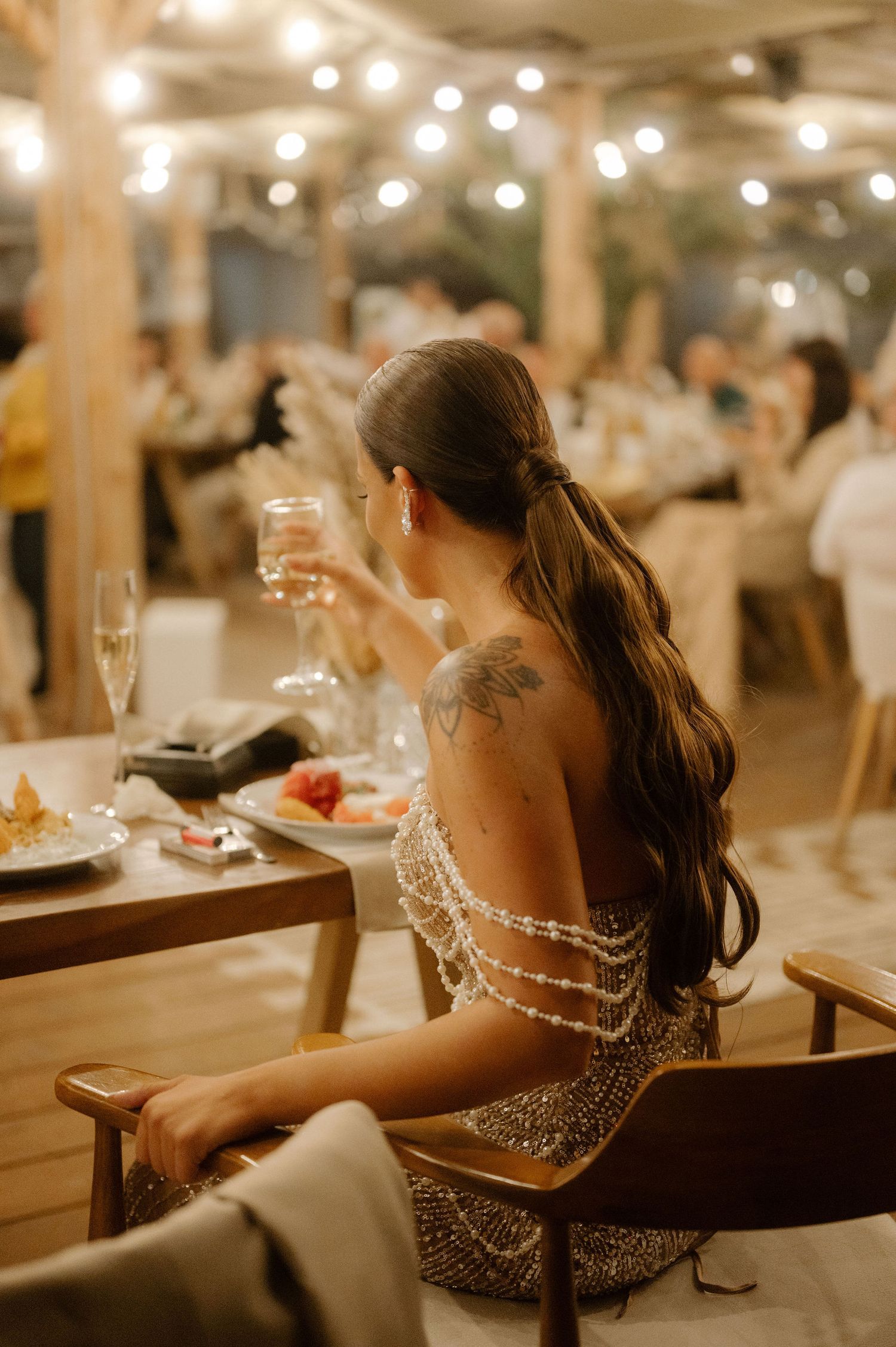 A guest in a sparkly dress with long dark hair sits at an elegantly decorated reception dinner table with string lights above.