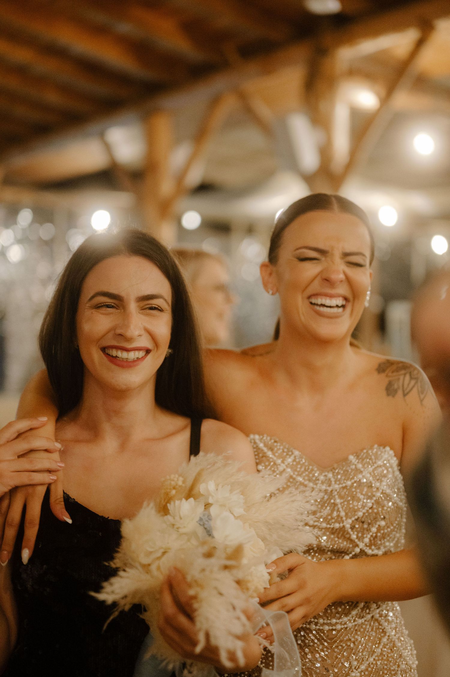 Two women share a joyful laugh at a wedding celebration with warm lighting and decorative elements in the background.