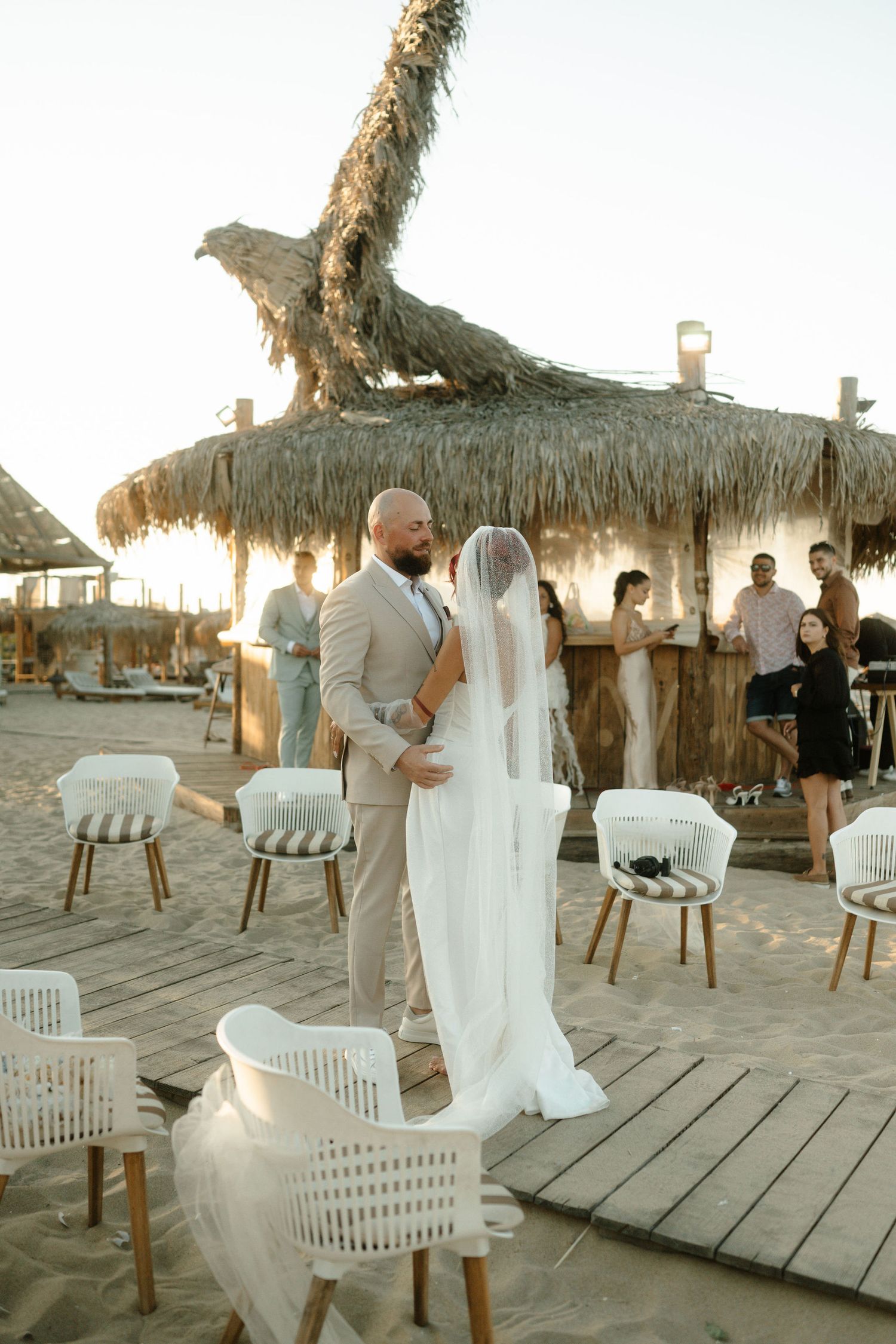 A romantic beach wedding ceremony setup with wooden deck, thatched umbrellas and white chairs at sunset.
