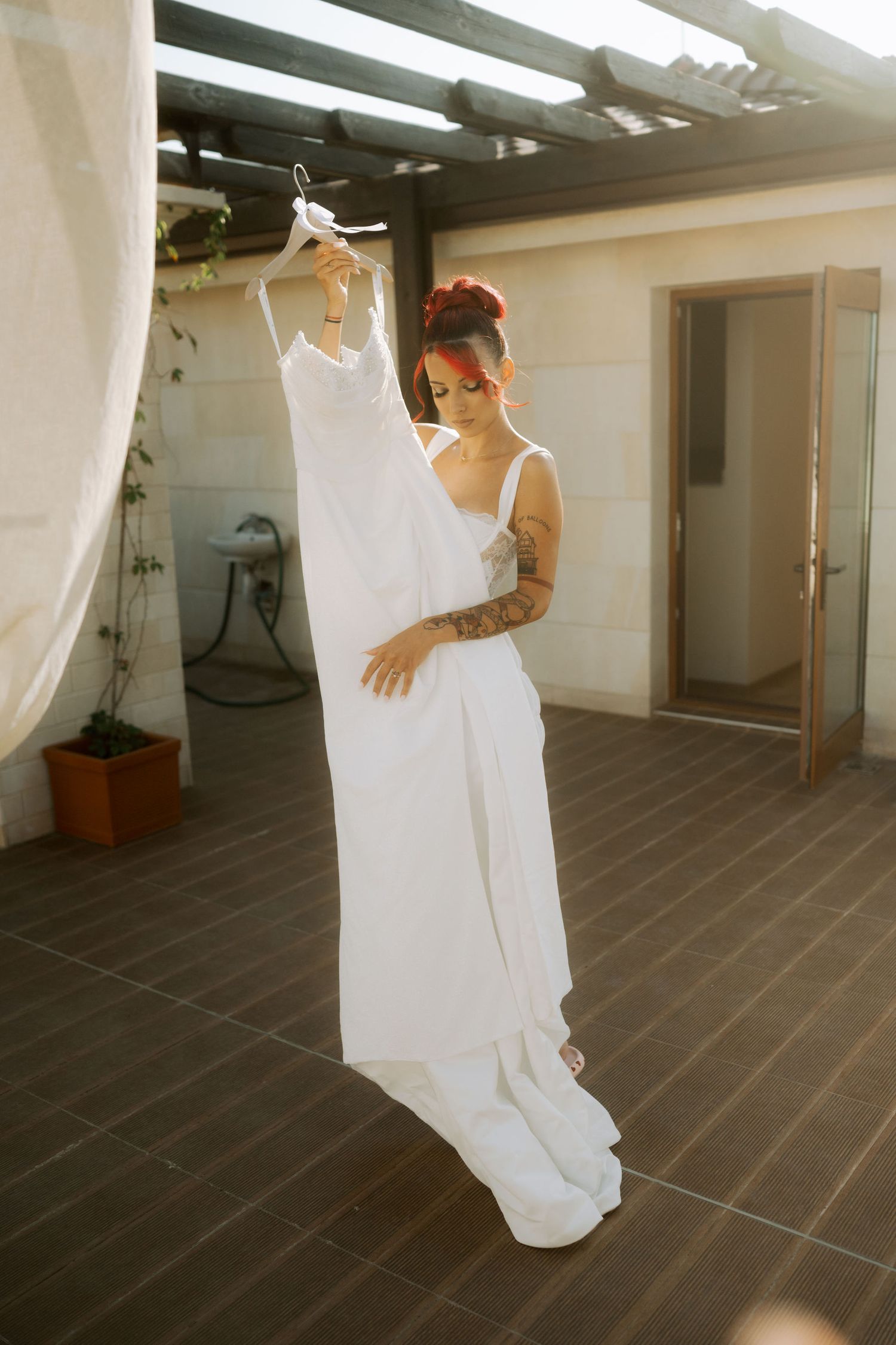 A person in a white wedding dress stands on a tiled patio while holding and examining the gown's flowing fabric.