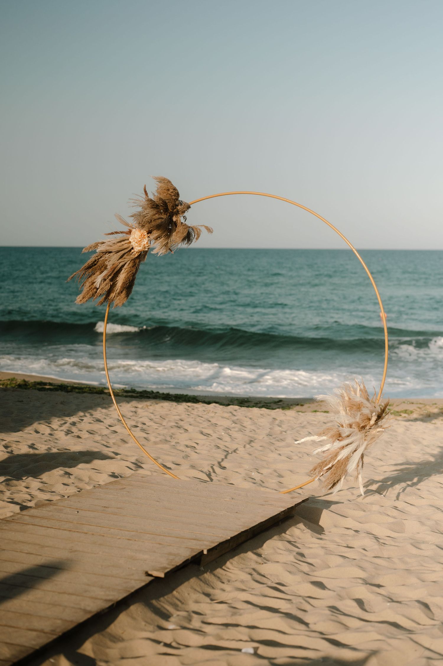 Wooden boardwalk path leads across sandy beach to ocean with dried palm fronds blowing in the wind at sunset.