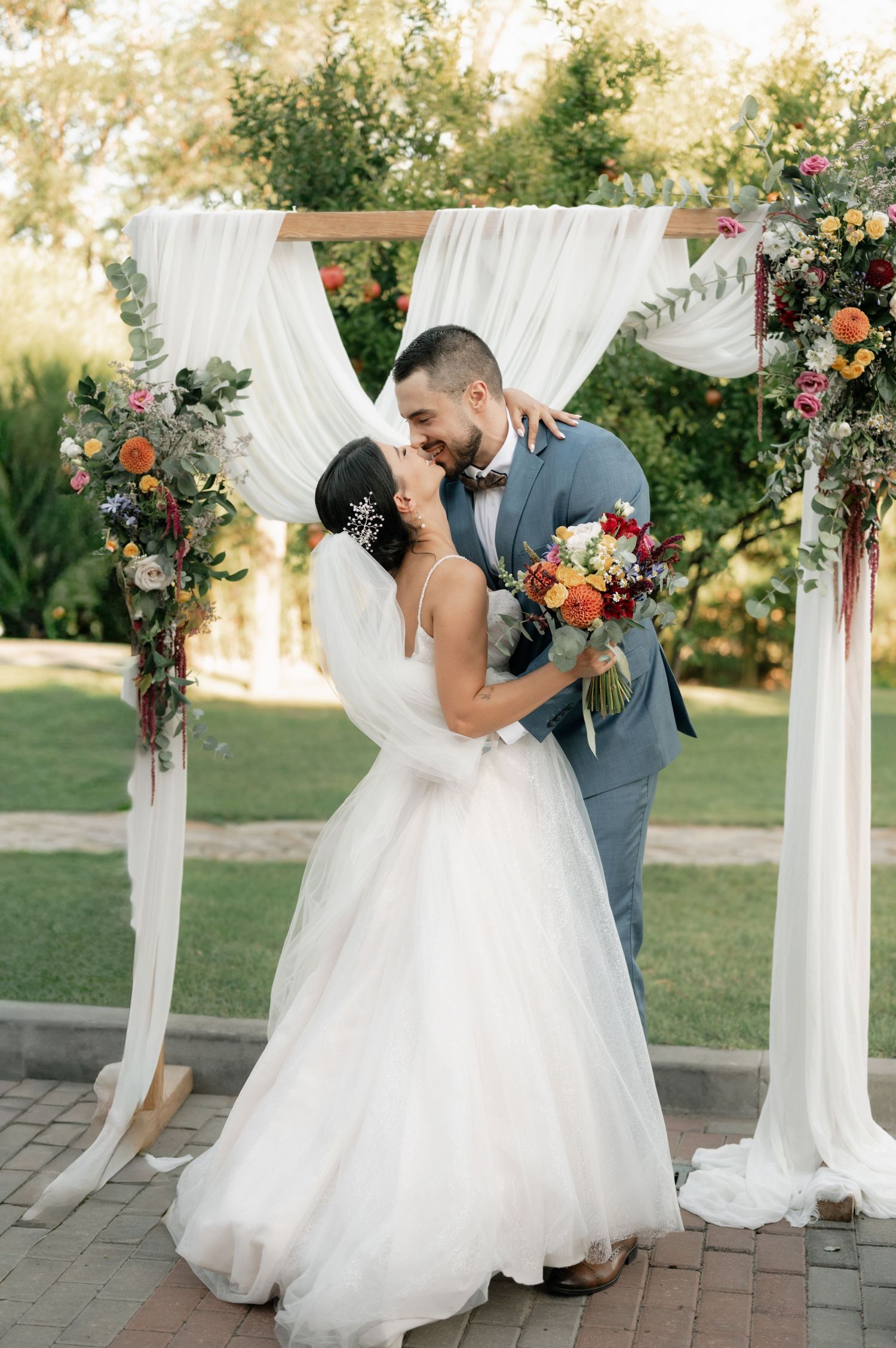 A romantic wedding moment under a white draped arch decorated with colorful florals on a brick patio at sunset.