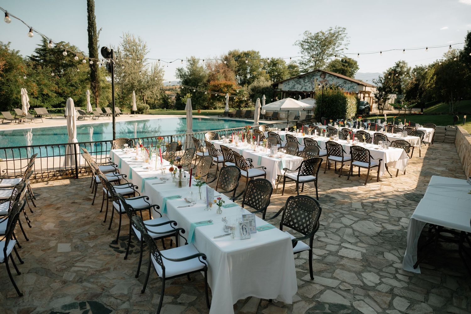 Elegant outdoor wedding reception setup with white-clothed tables and chairs arranged on a stone patio near a pool.