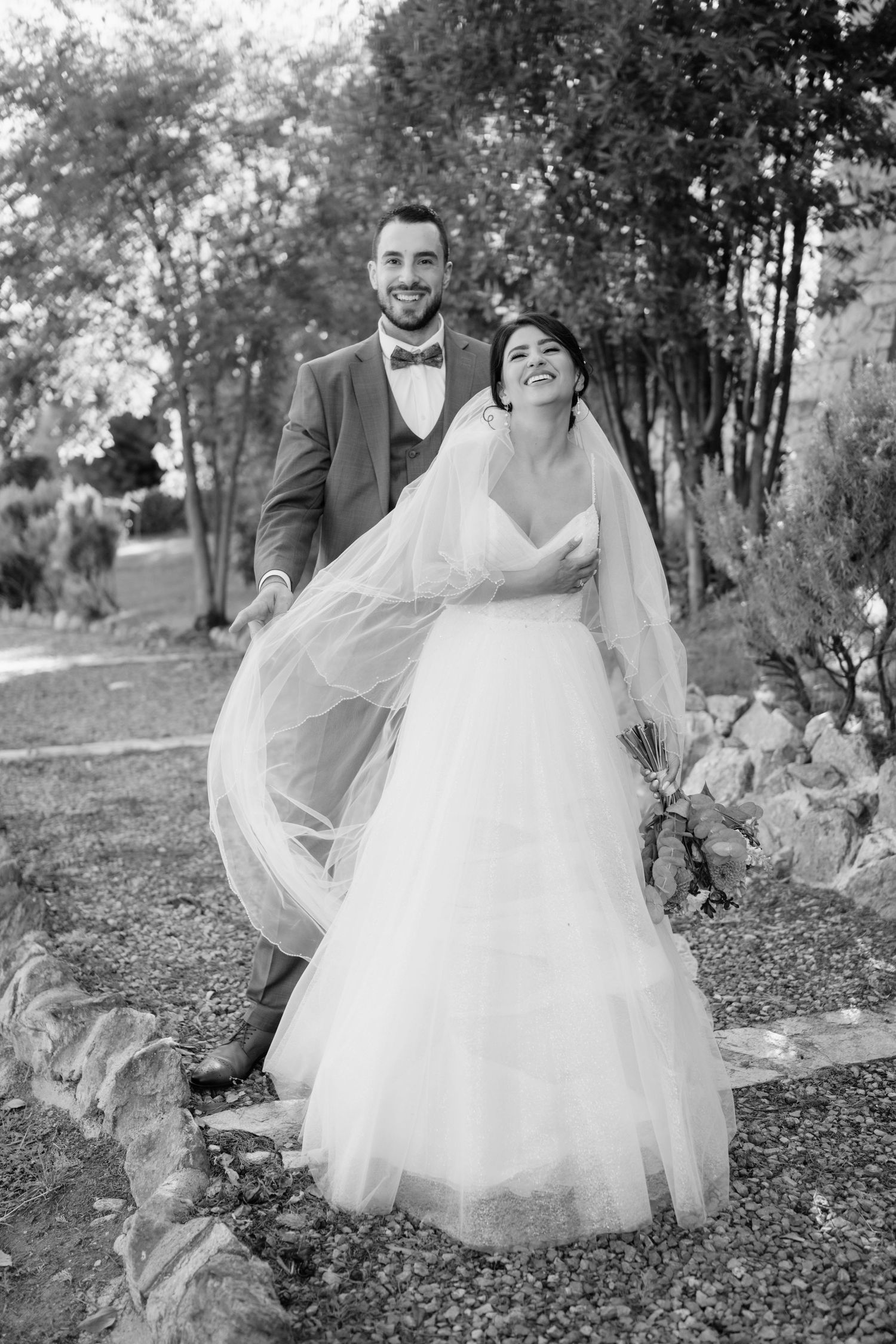 A couple in wedding attire poses together on a scenic outdoor path surrounded by trees and foliage.
