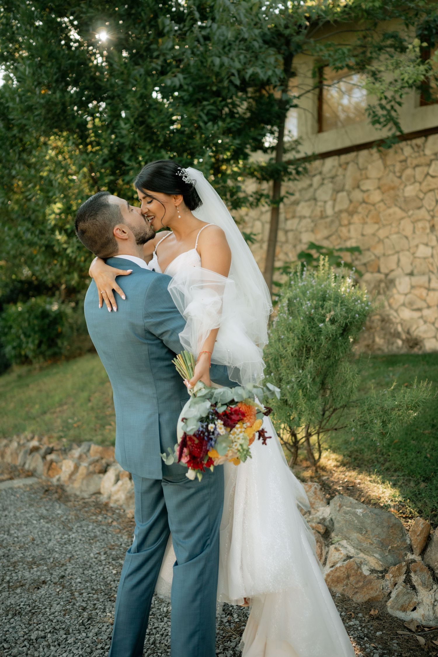 A romantic wedding couple embraces on a gravel path beside a rustic stone wall during golden hour.
