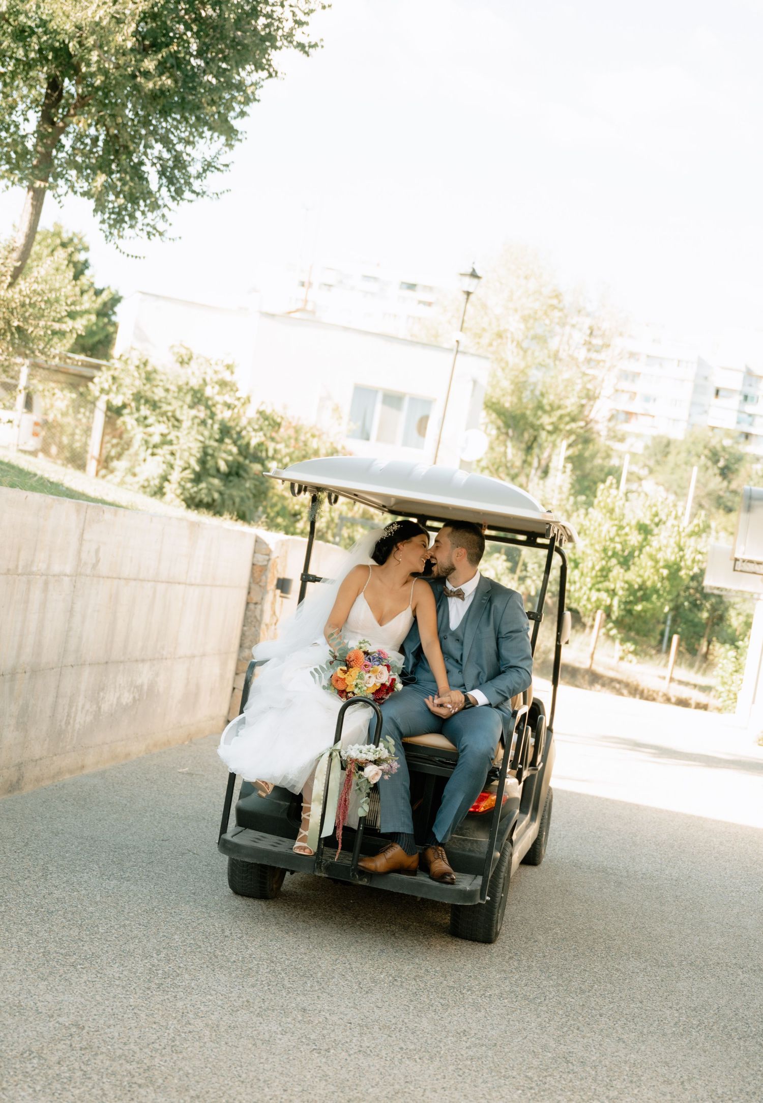 A couple in formal attire rides away in a golf cart through a sunny outdoor venue after their wedding celebration.