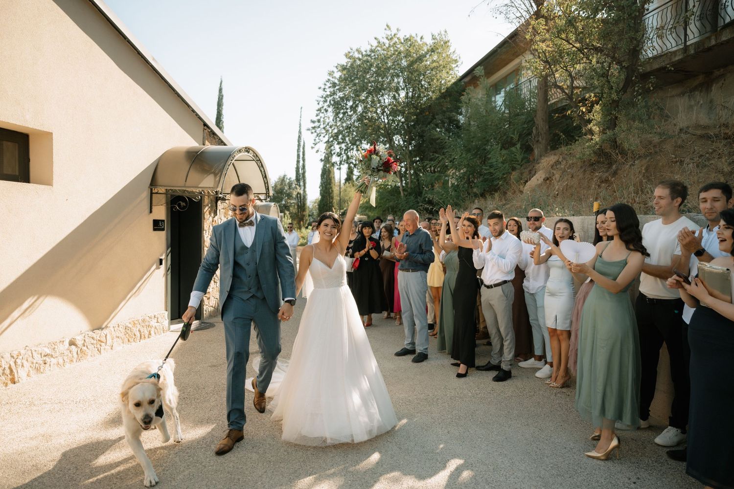 Newlywed couple walks down path with small dog while wedding guests look on during outdoor summer celebration.