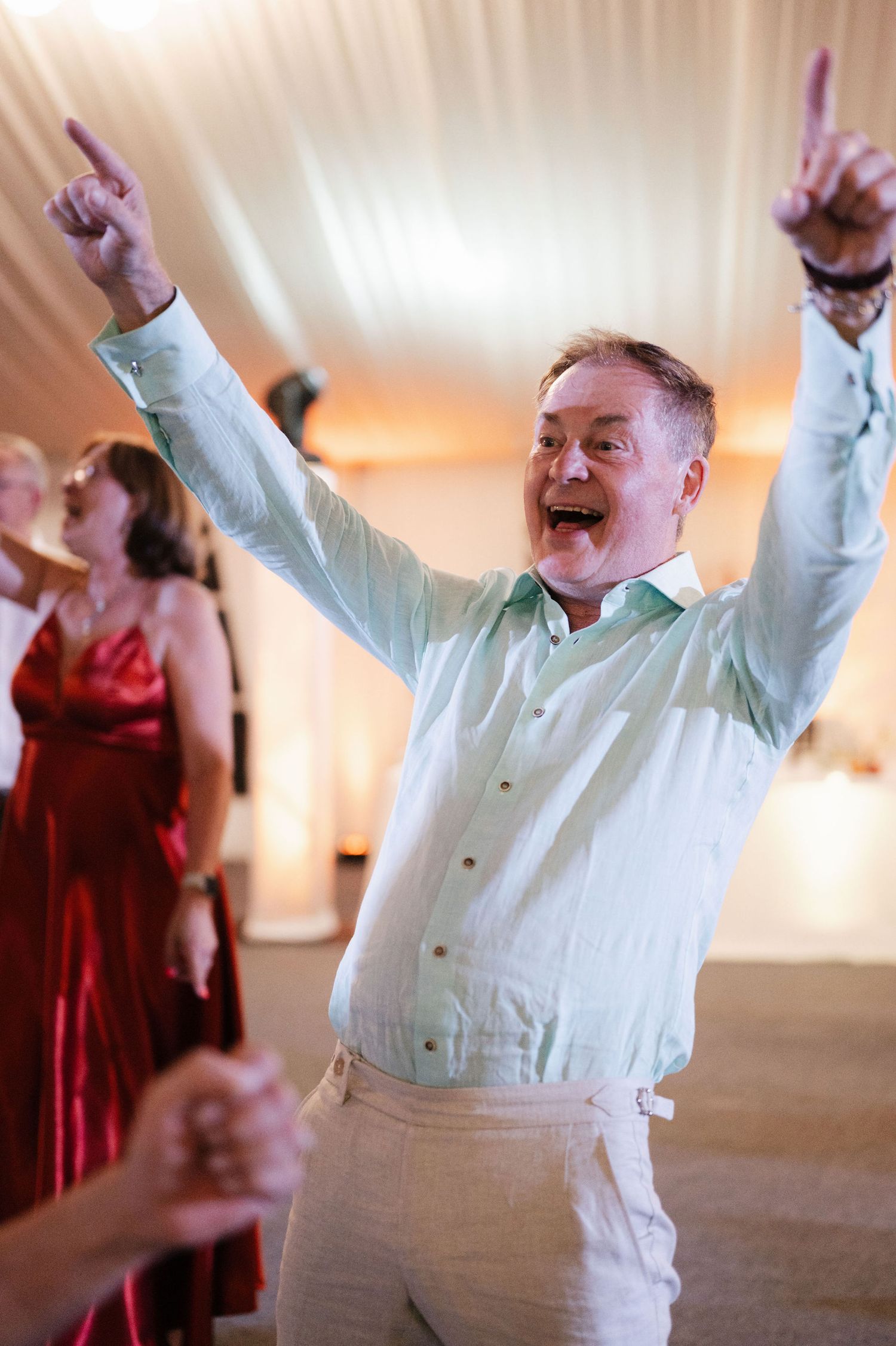 A person in a white dress shirt dances enthusiastically at a wedding reception with arms raised in celebration.