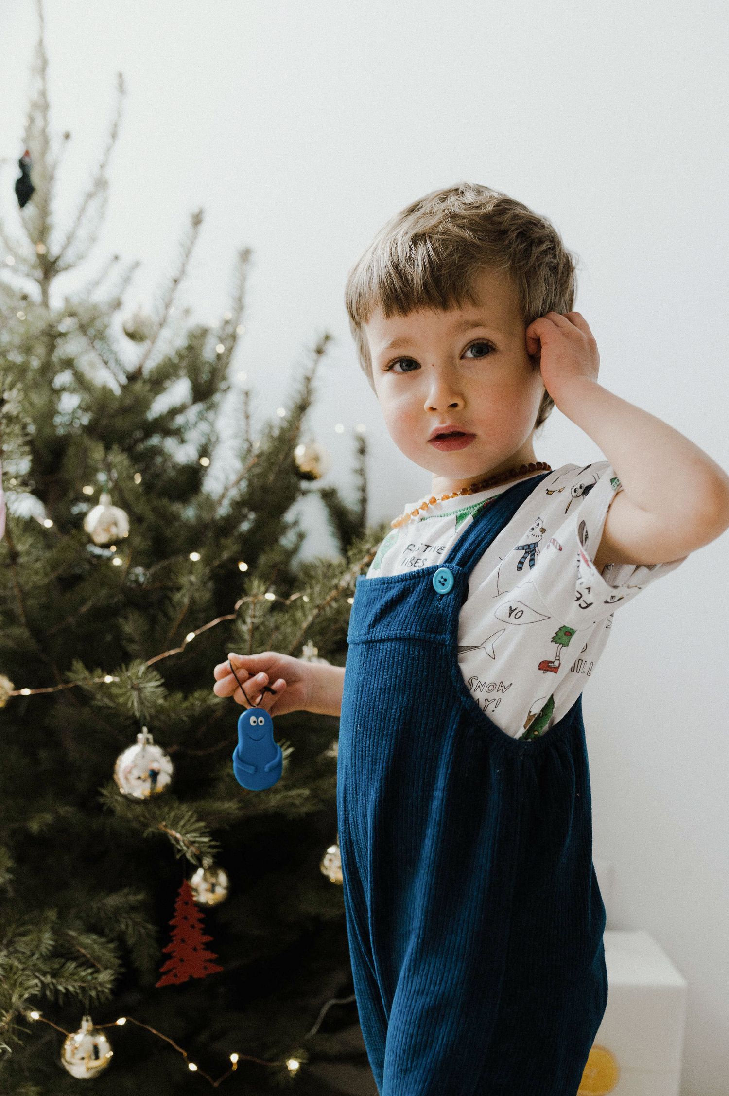 A toddler in a blue dress and white collar plays near evergreen trees and holiday decorations.