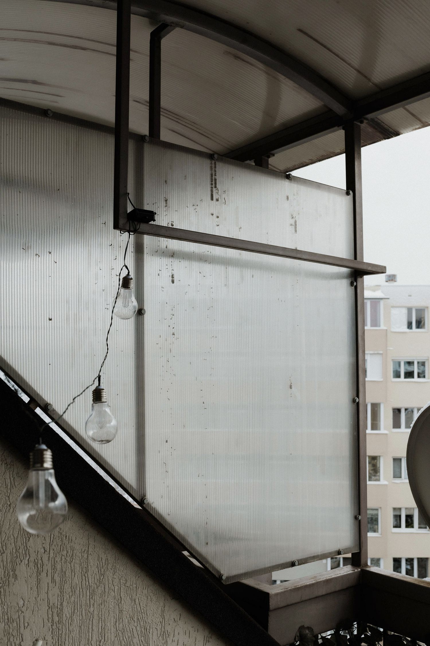 Light bulbs hang from a metal bar on a weathered white balcony with an apartment building visible in the background.