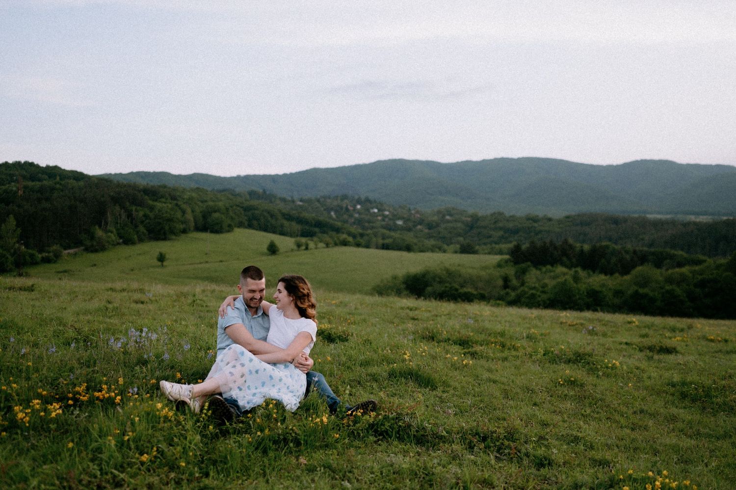 A couple embraces on a hilltop meadow overlooking a mountainous landscape.