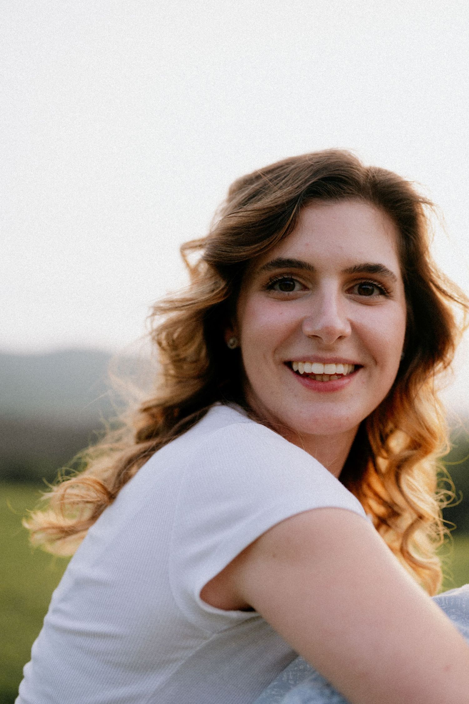 A person with flowing hair smiles warmly while posing outdoors in natural light.