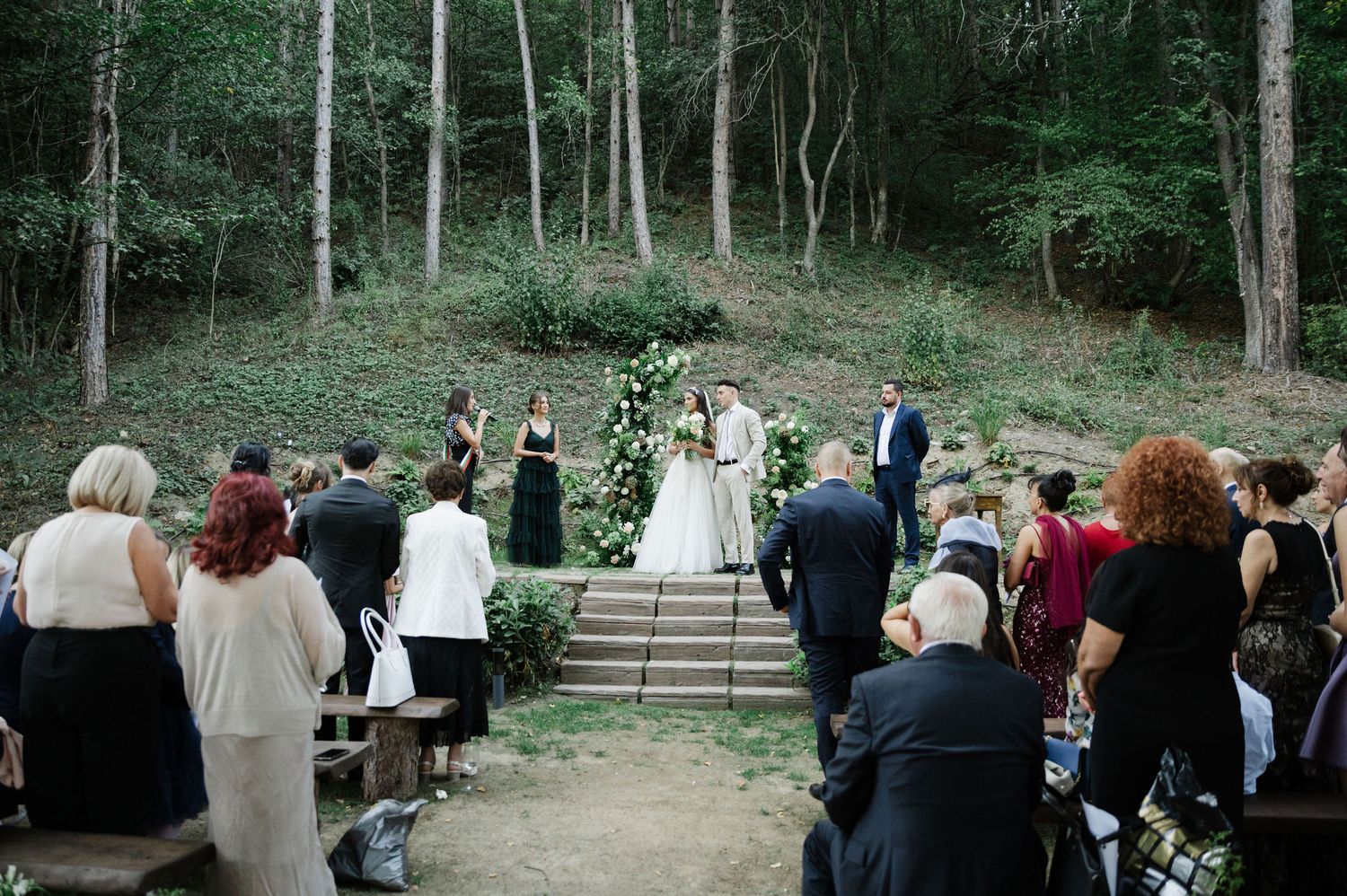 Wedding guests seated on hillside watching ceremony on stone steps in forest setting.