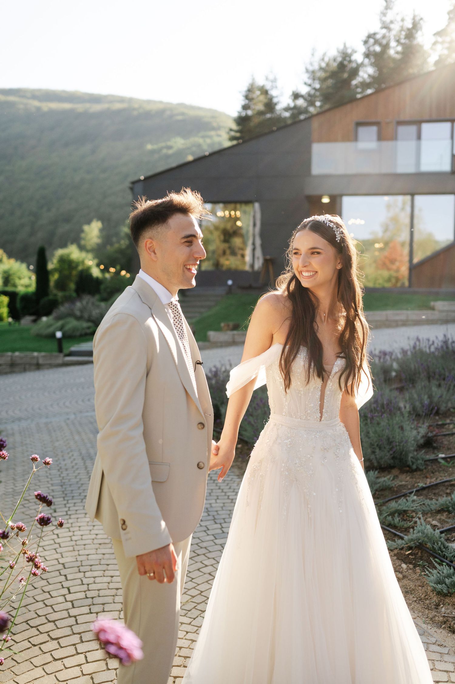 Newlyweds laugh together on a stone patio surrounded by pink flowers and mountains at golden hour.