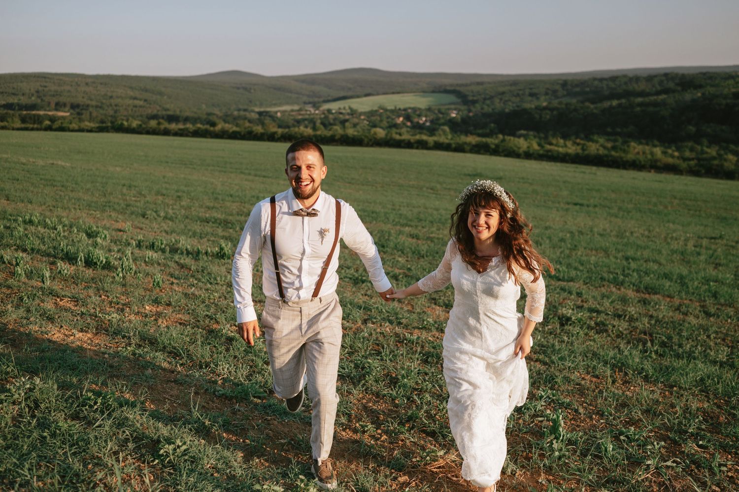 A couple in white wedding attire runs playfully through a green hillside meadow at sunset.