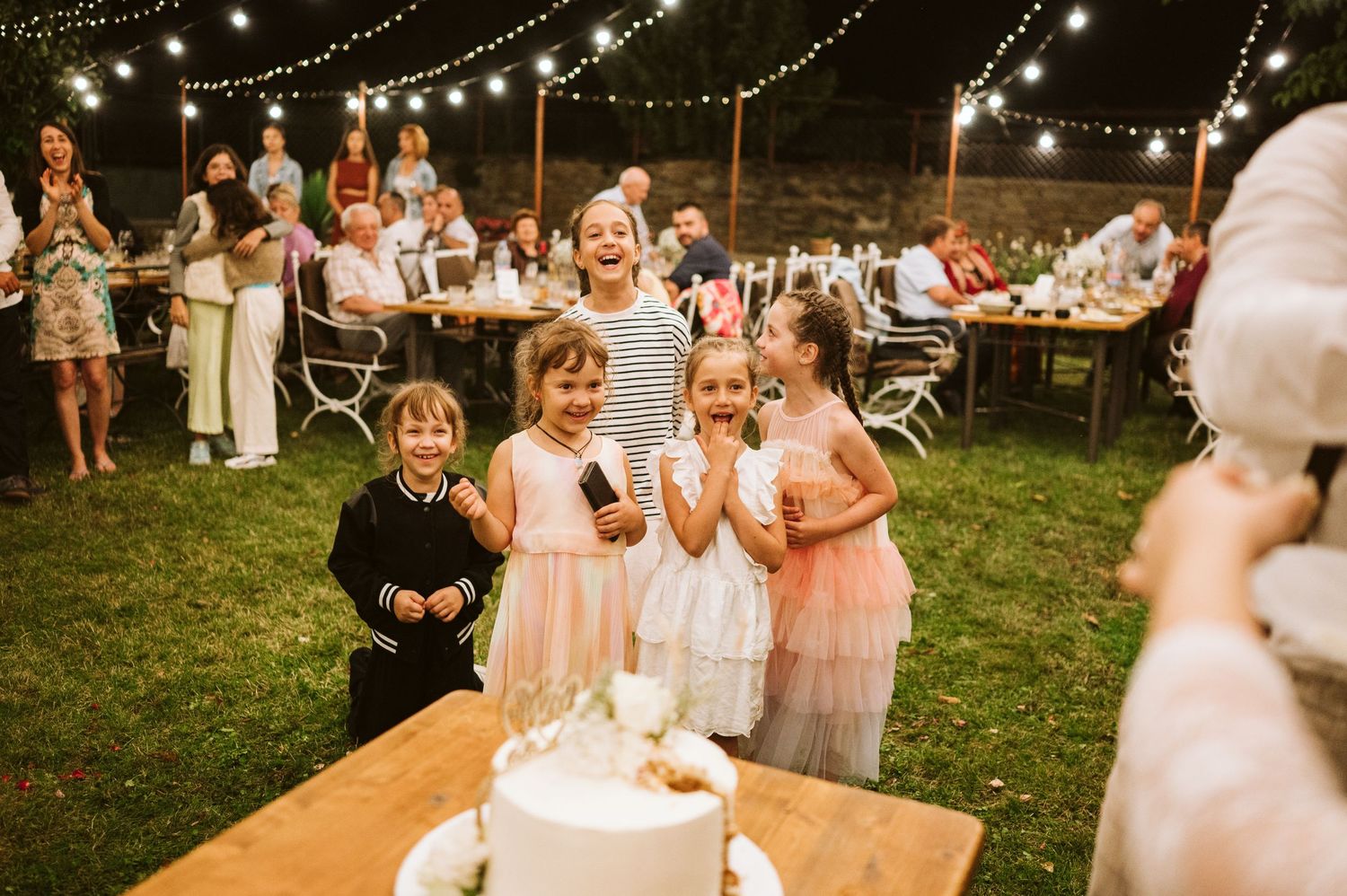 Children in pastel dresses gather around a white wedding cake at an outdoor reception.