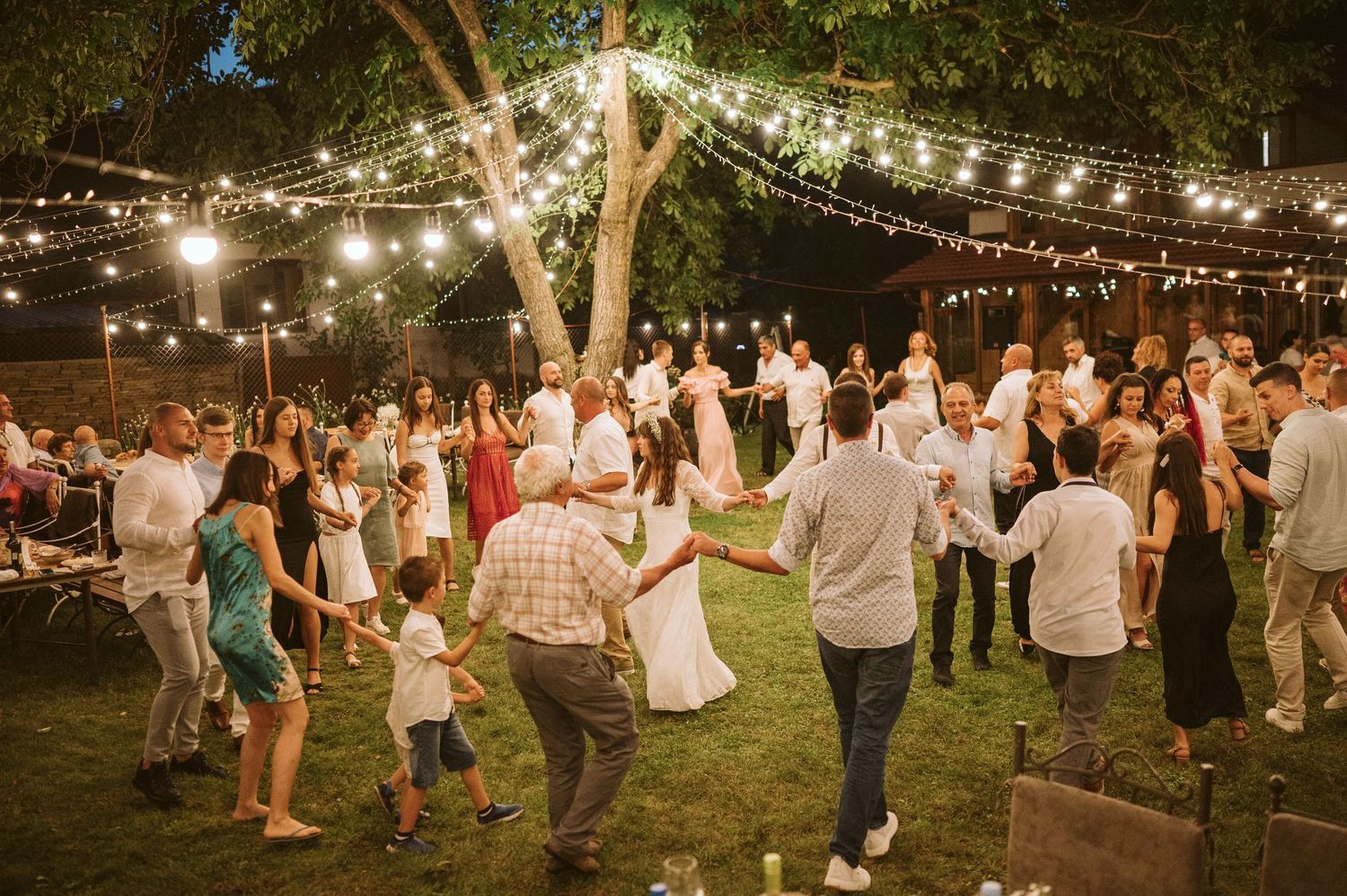 Wedding guests dancing and celebrating on a lawn under warm string lights at night.
