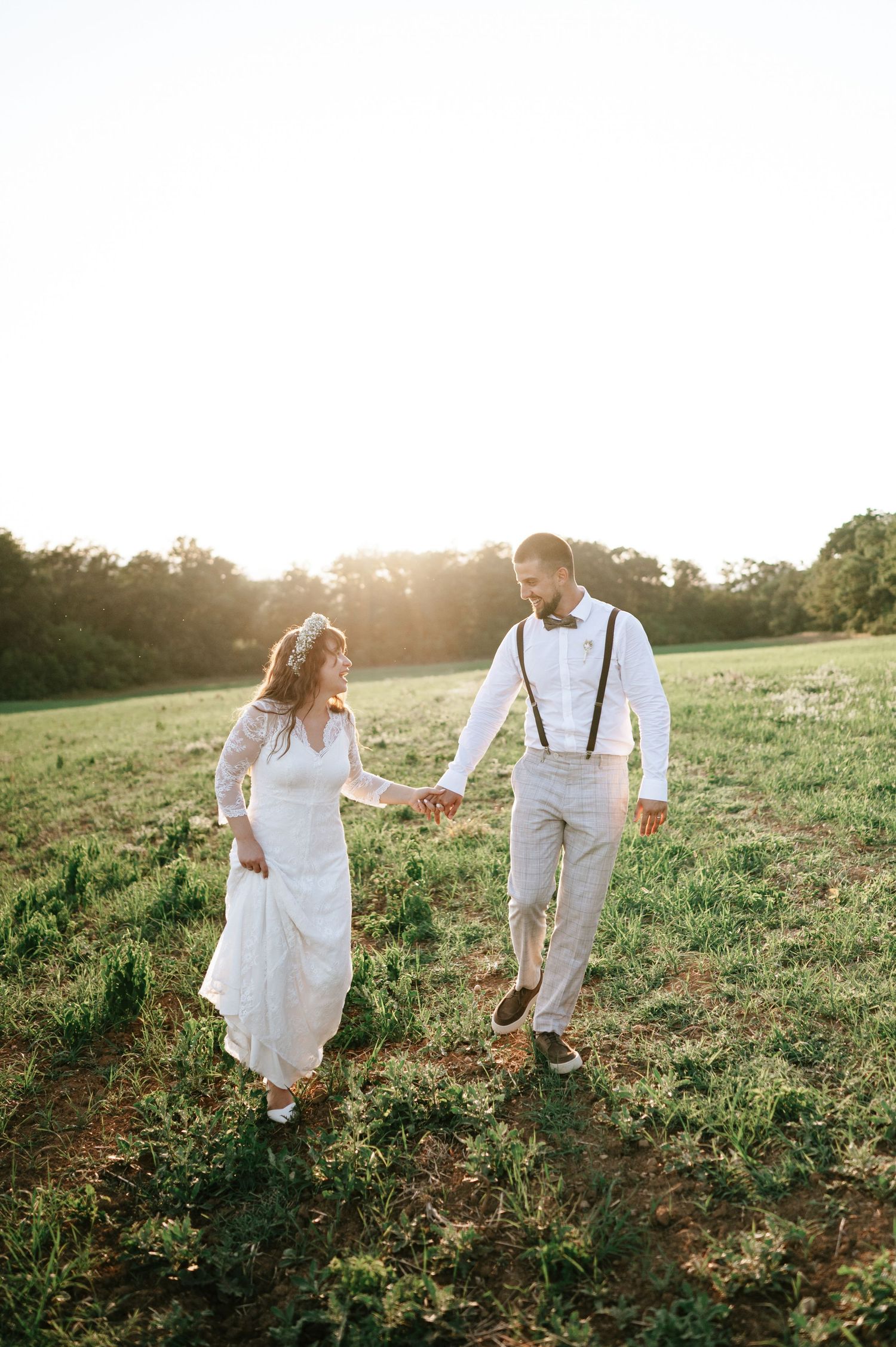 Couple strolling through a grassy field at sunset in elegant wedding day outfits.